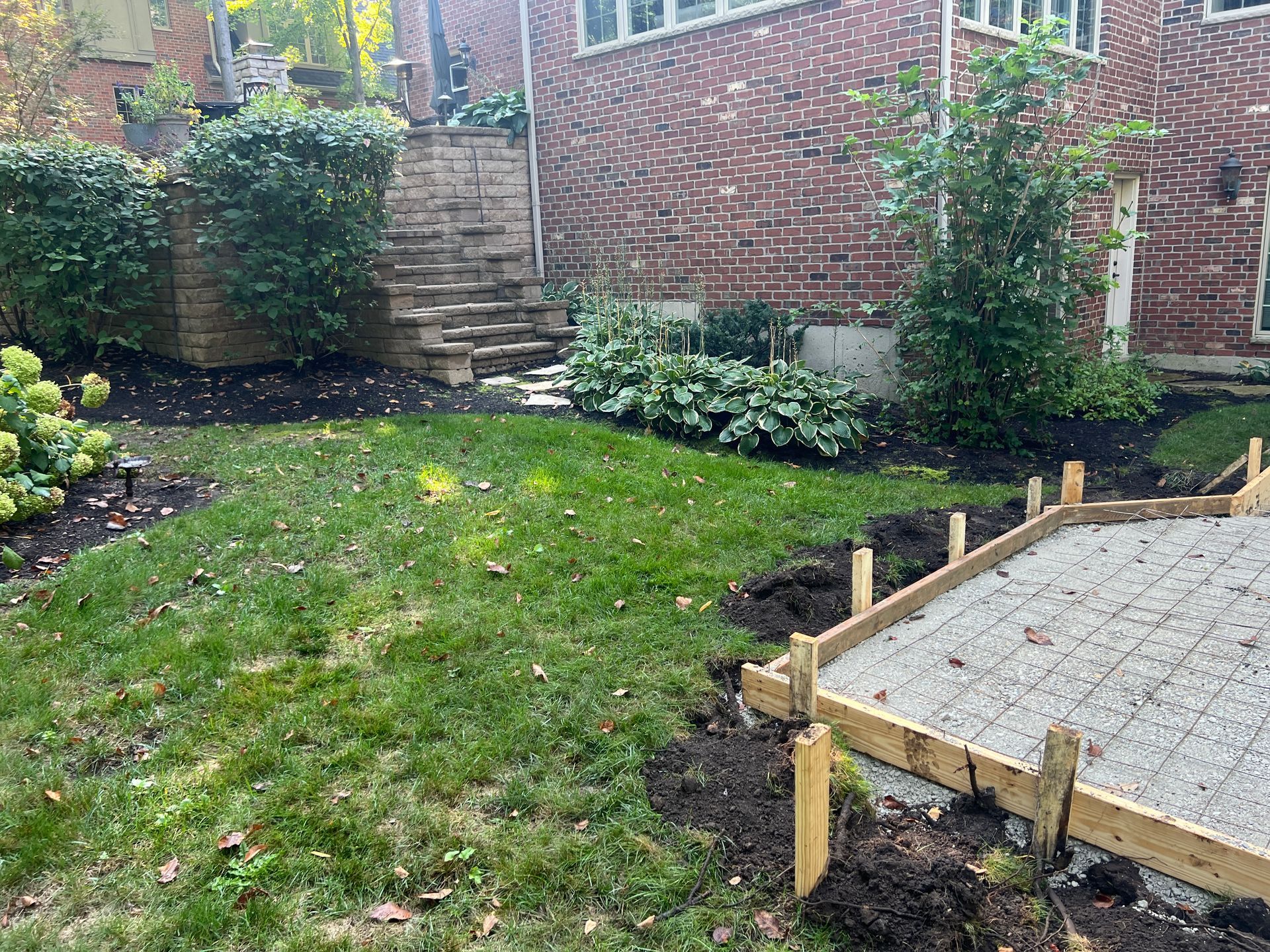 A wooden frame borders a gravel patio foundation in a backyard, with a brick house and landscaped garden in the background.