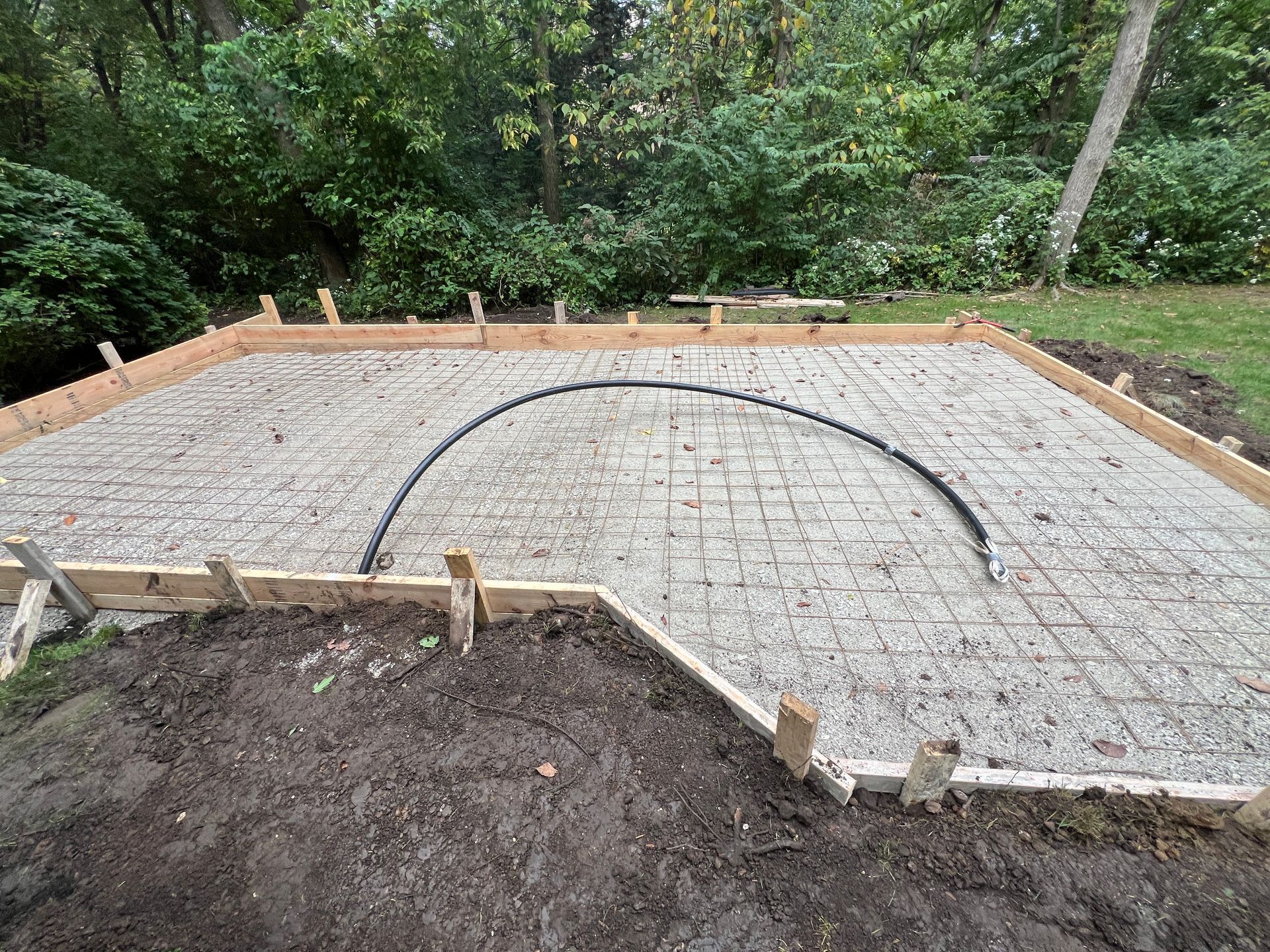 A rectangular dirt area prepped for a concrete pour, framed by wooden boards with a wire mesh grid and a curved conduit.