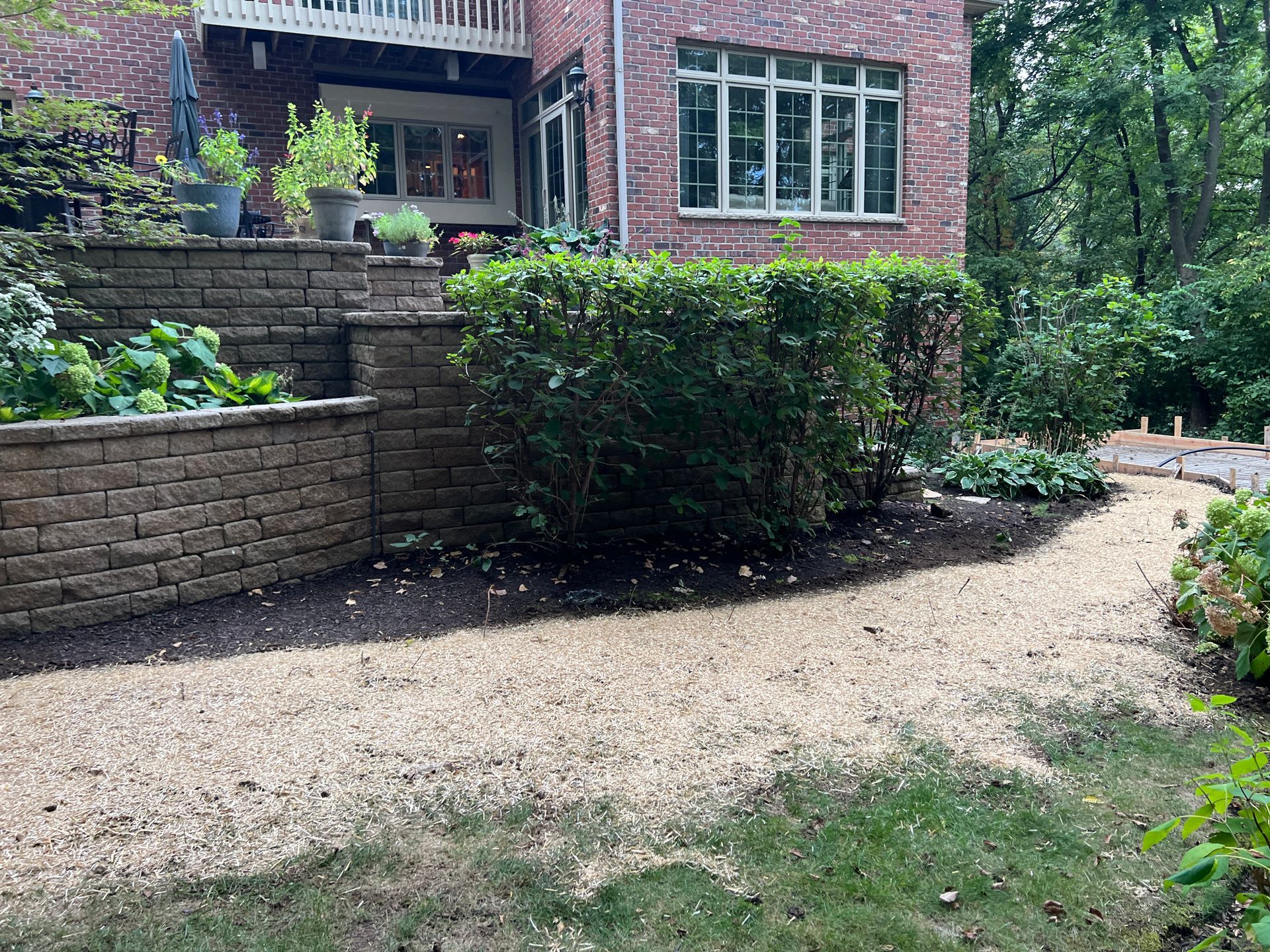 A brick house with a tiered stone retaining wall, lush green shrubs, and a gravel path leading through the garden.