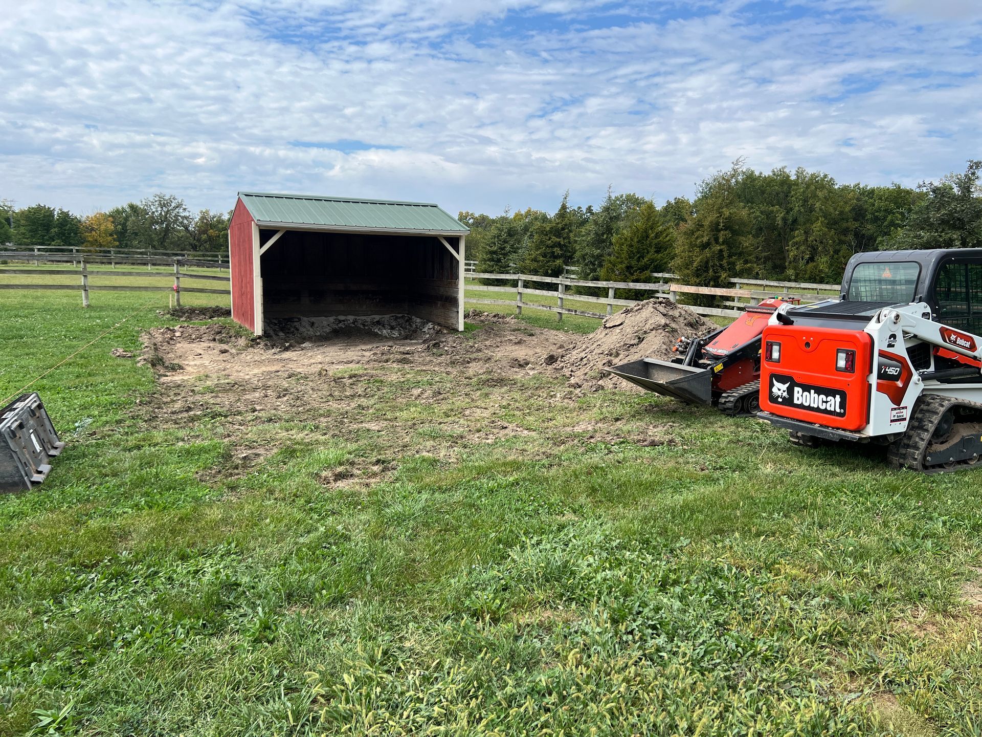 A red and green shed sits in a grassy field next to a red Bobcat skid-steer loader during a landscaping project.