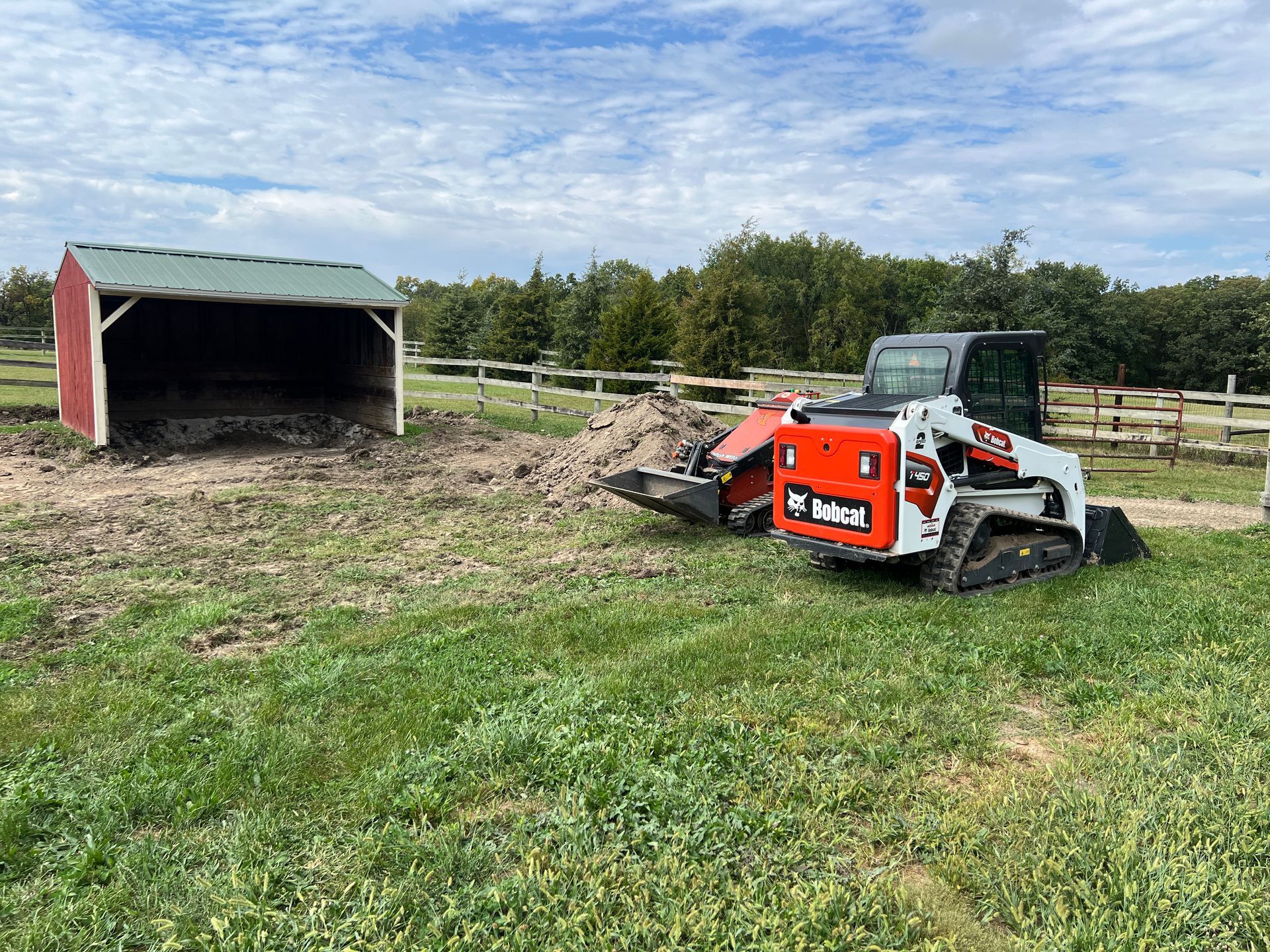 A Bobcat track loader parked in a grassy field next to a small red shed and a pile of dirt under a partly cloudy sky.