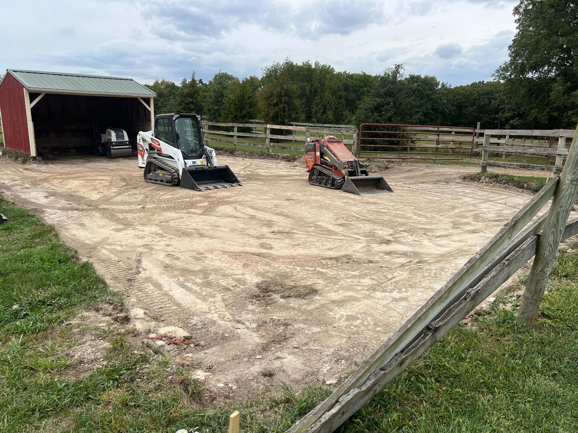 A white skid steer and a small orange excavator sit on a dirt lot next to a red shed in a rural, fenced field.