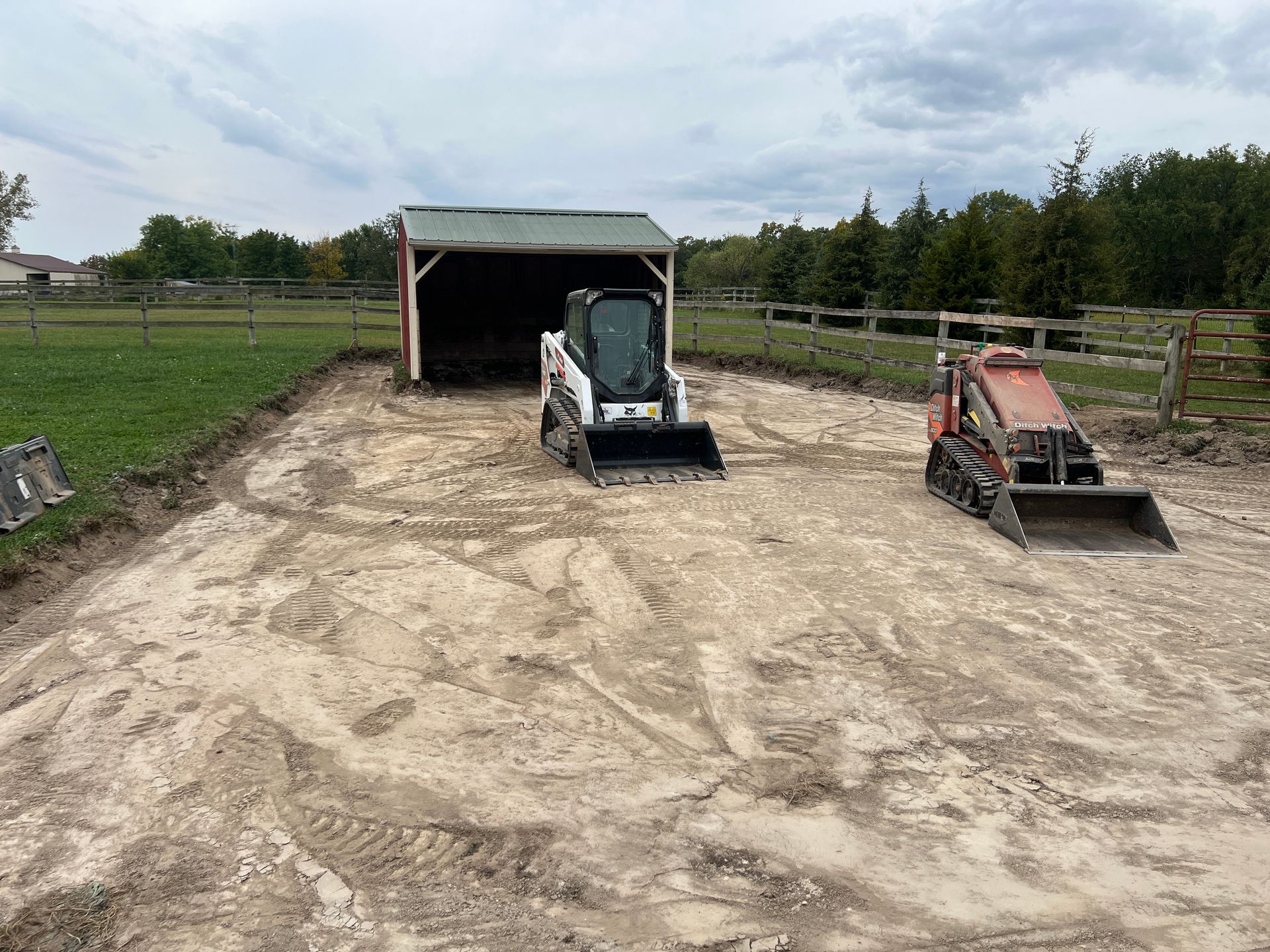 A white Bobcat skid-steer and a smaller orange tracked loader on a cleared, sandy construction site in front of a shed.