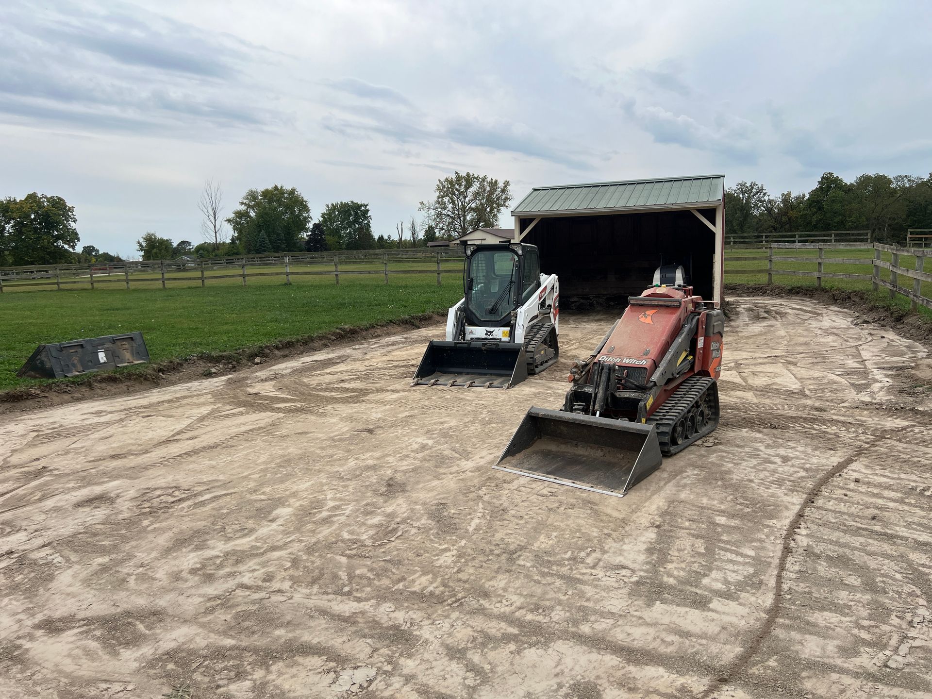 A white skid steer and an orange tracked loader parked on a dirt lot in front of a shed in a grassy field.