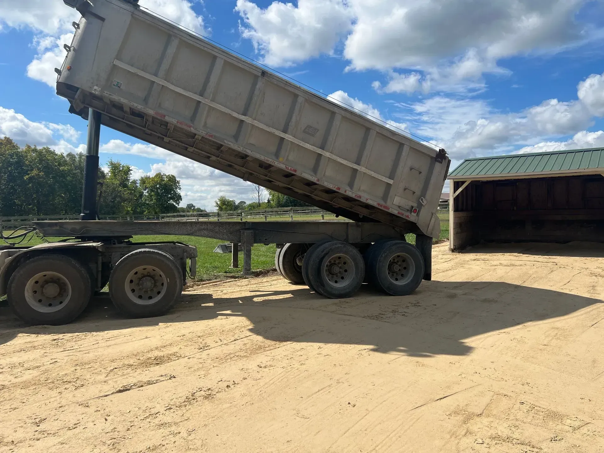 A gray dump trailer in a raised, unloaded position parked on a sandy surface near a shed under a partly cloudy blue sky.