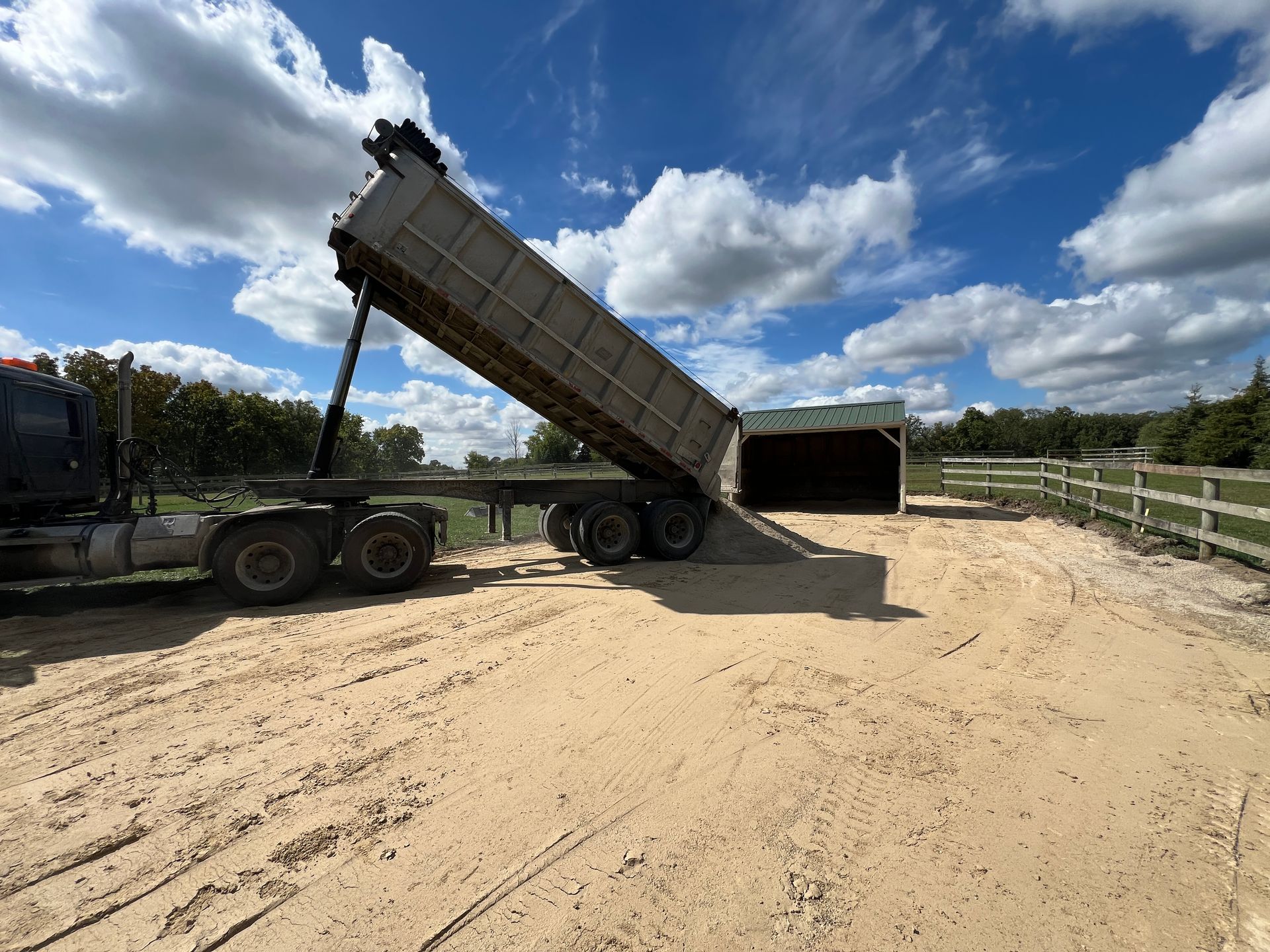 A large dump truck in a sandy lot is raised to unload material near a shed under a bright, partly cloudy blue sky.
