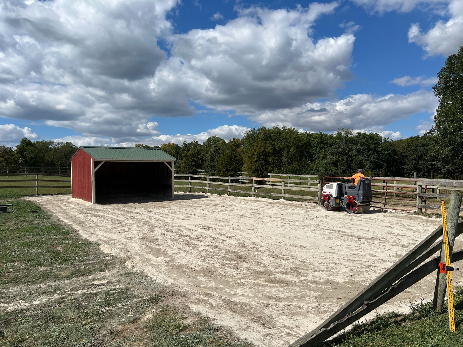 A person operates a small construction vehicle on a light-colored dirt lot next to a red and green shed.