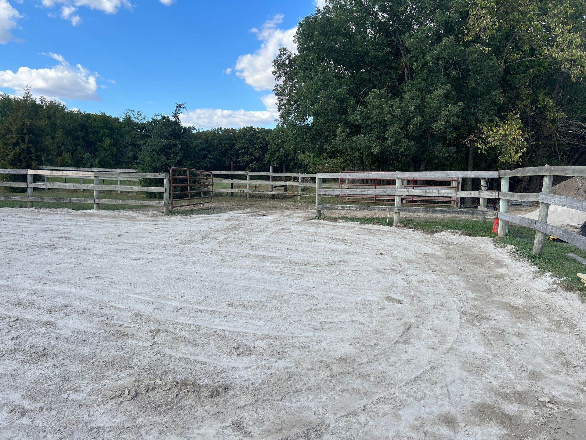 A light-colored, circular riding arena with white sand, enclosed by wooden fencing and surrounded by dense green trees.