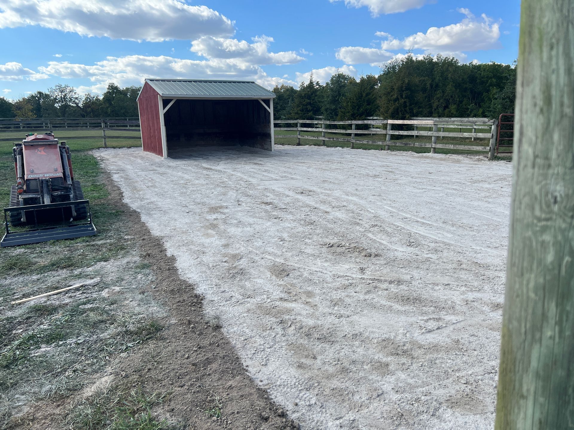 A red-and-white animal shelter sits on a white gravel pad in a fenced field under a cloudy sky, with machinery nearby.
