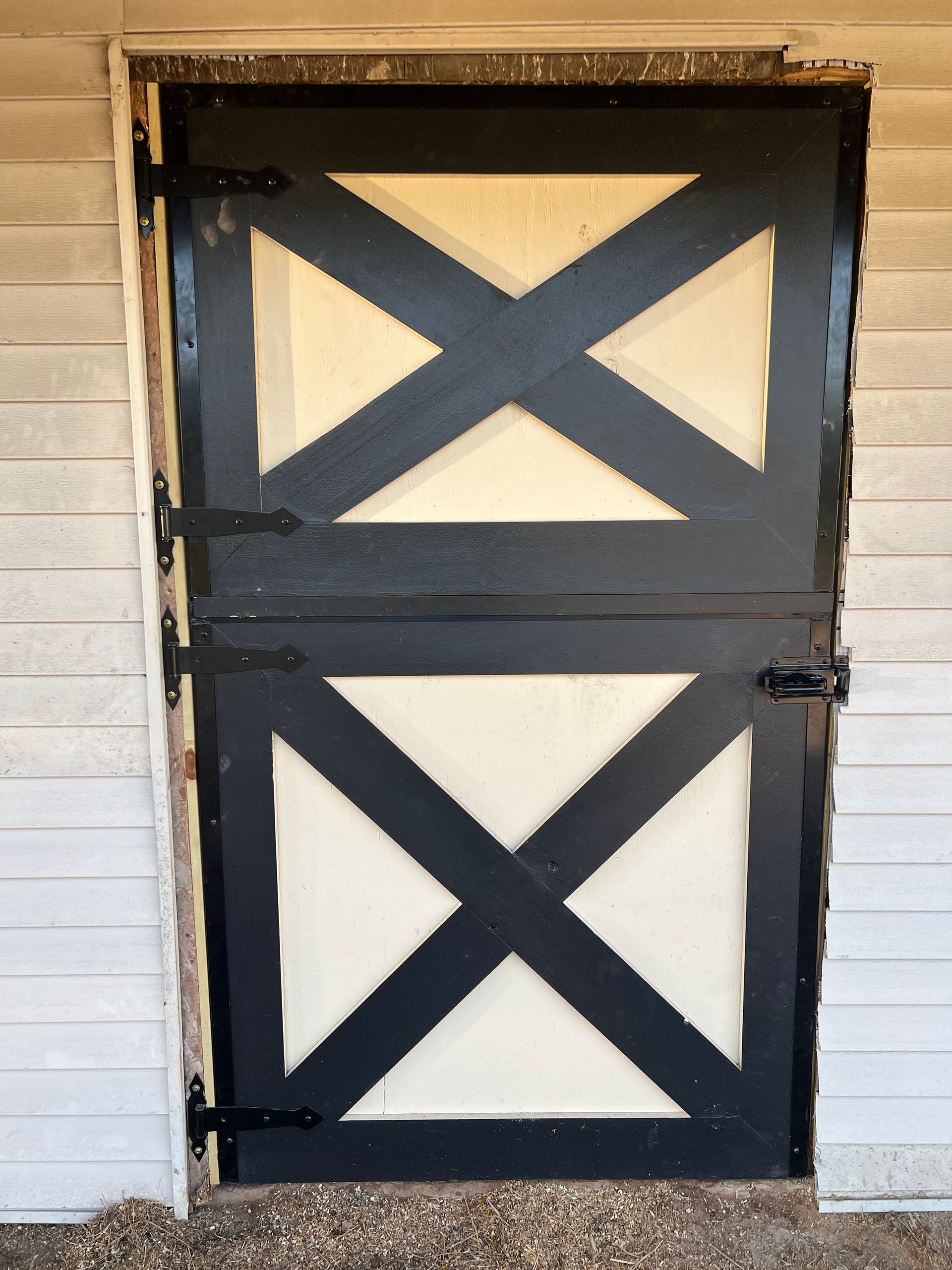 A black-trimmed, white-paneled Dutch stable door with an X-brace design, set in a light grey horizontal-siding wall.