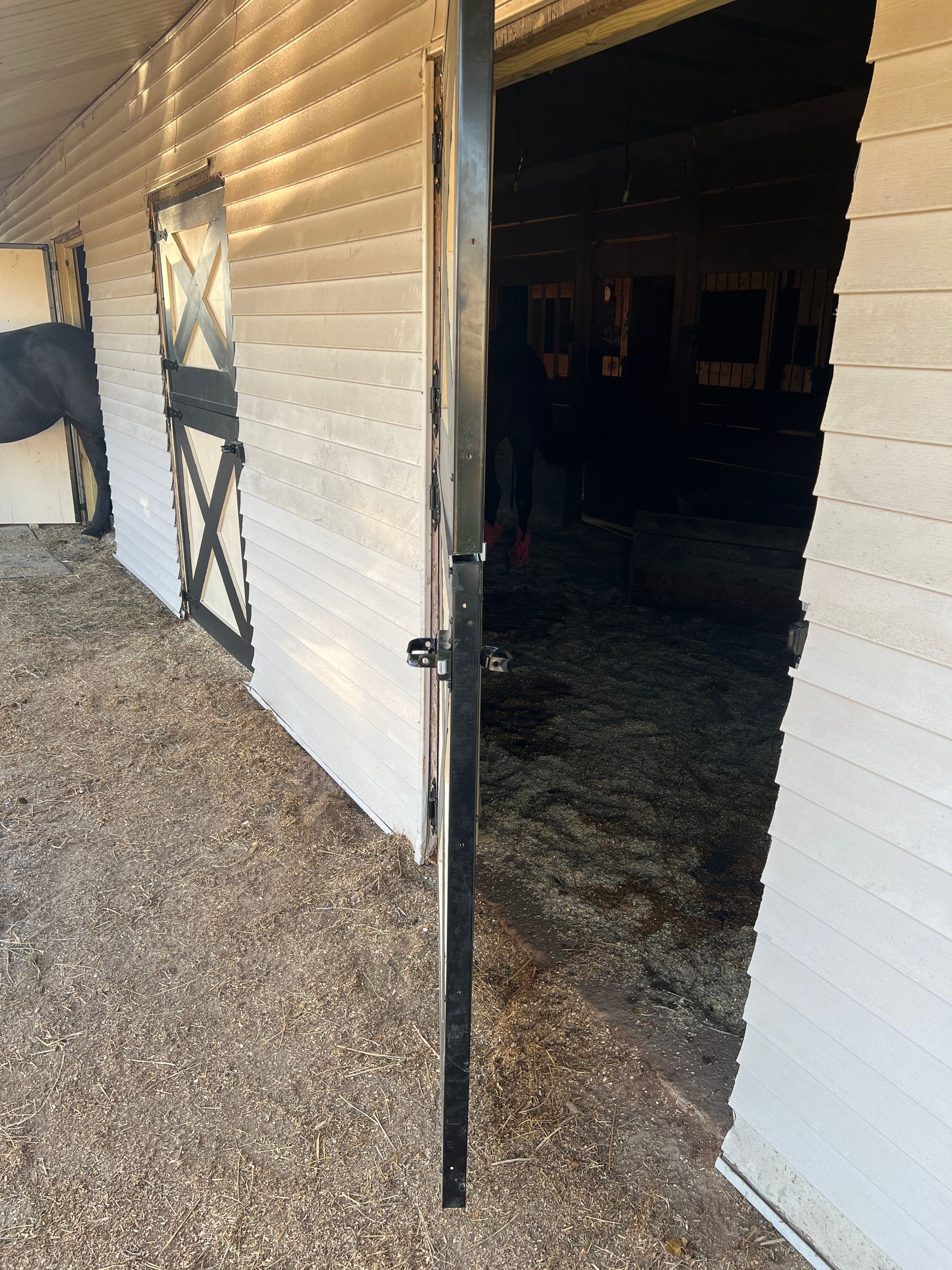 A horse stall doorway with white siding, showing a partially open black metal gate and a dark interior with bedding.