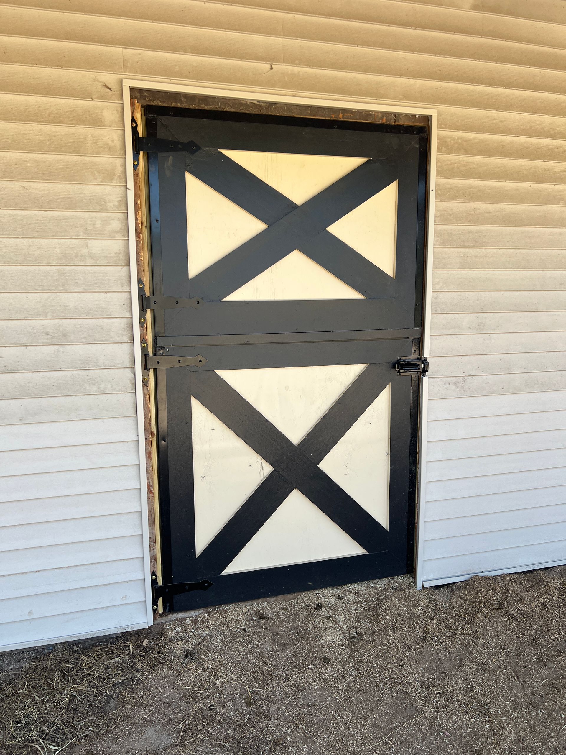 A black and cream-colored Dutch-style barn door with an X-pattern design, set into a white siding wall.
