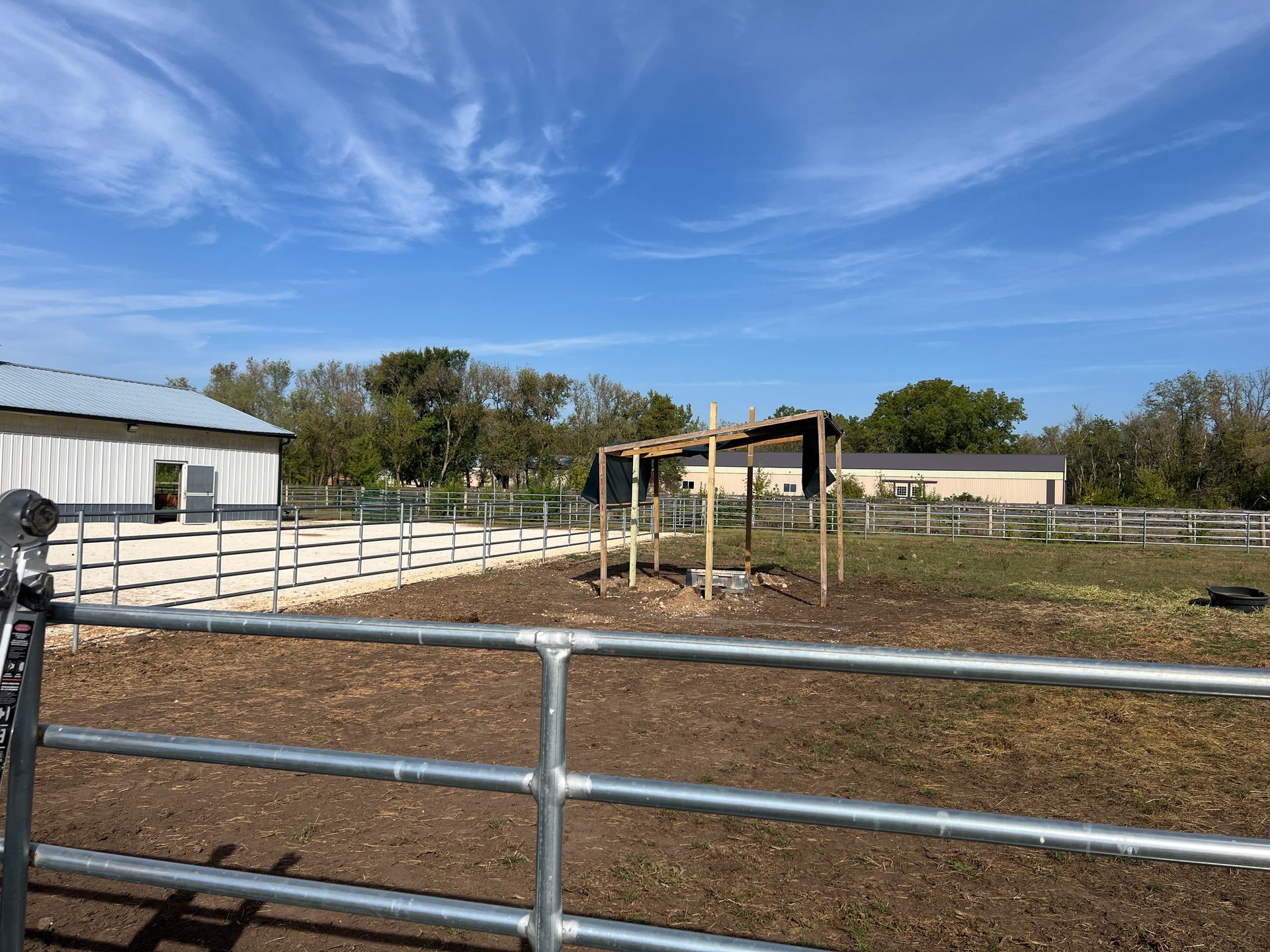 A fenced-in dirt pasture with a small wooden shelter under a bright blue, partially cloudy sky.