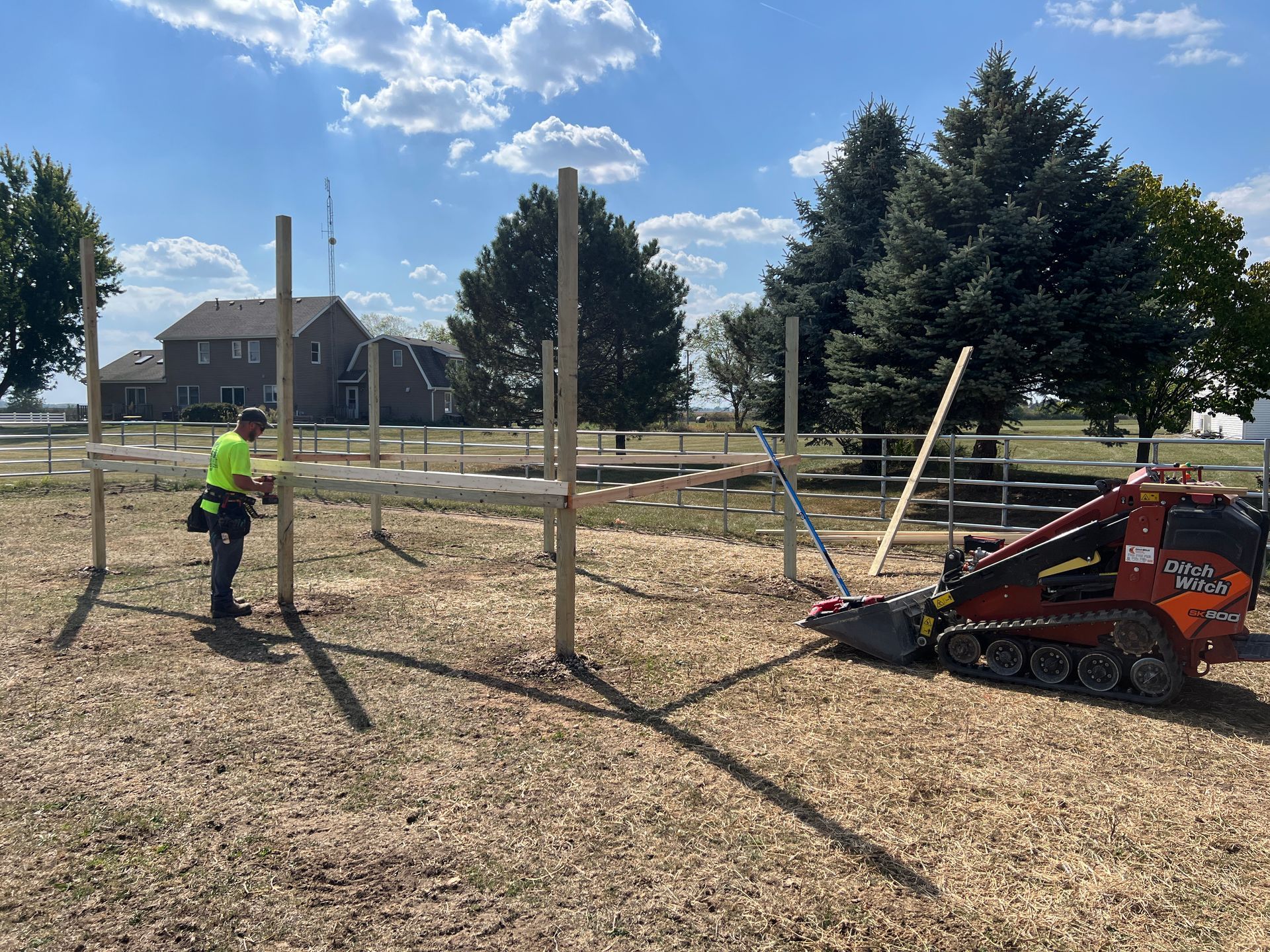 A worker installs wooden posts for a structure on a sunny day with a skid steer loader parked nearby.