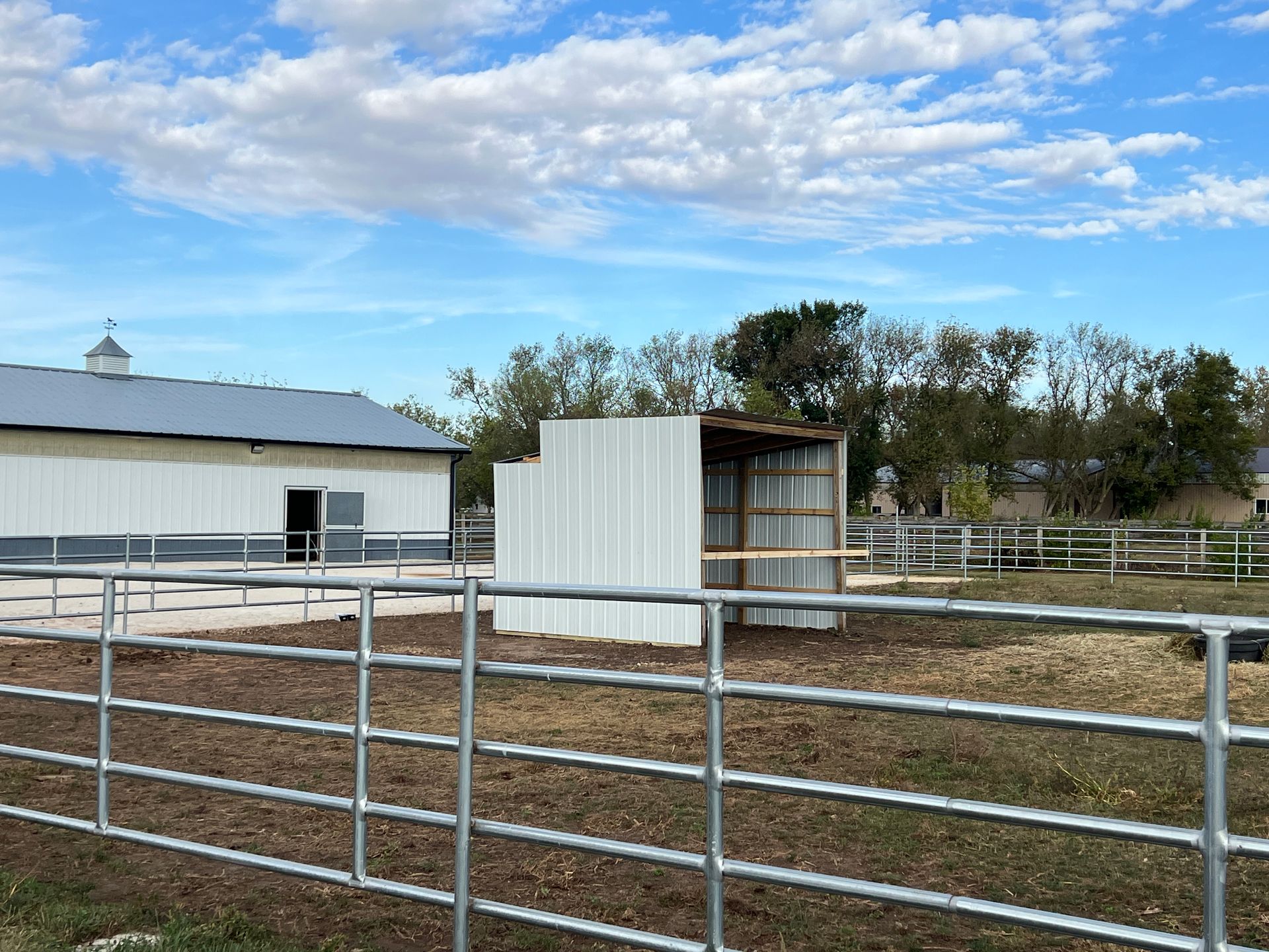 A white metal barn and run-in shed stand in a fenced dirt paddock under a blue sky with scattered white clouds.