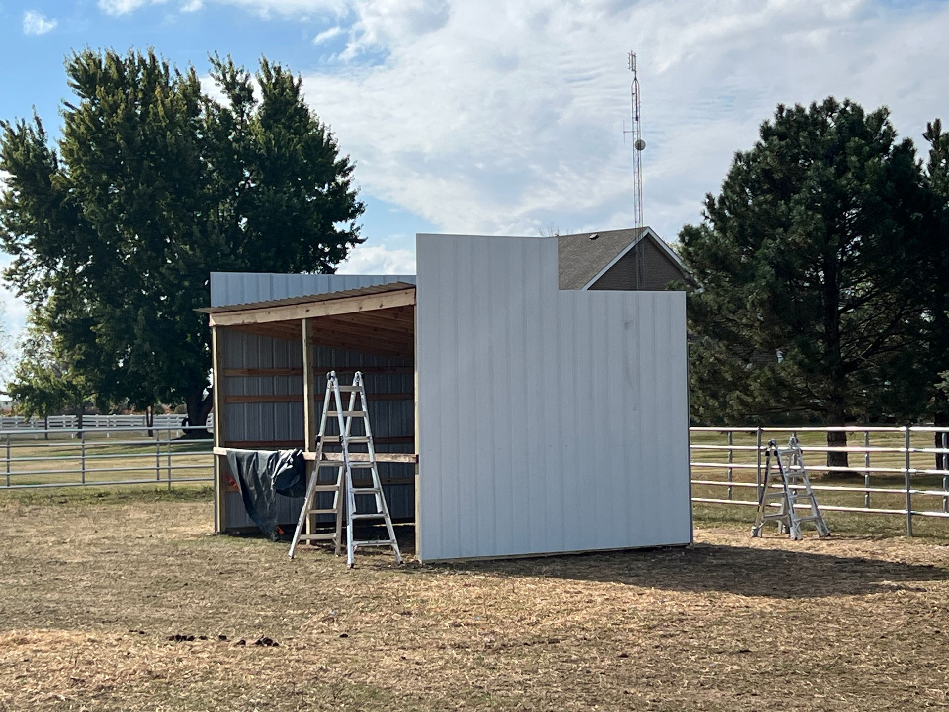 A partial metal structure under construction in a grassy field, featuring white siding, an open side, and a ladder.
