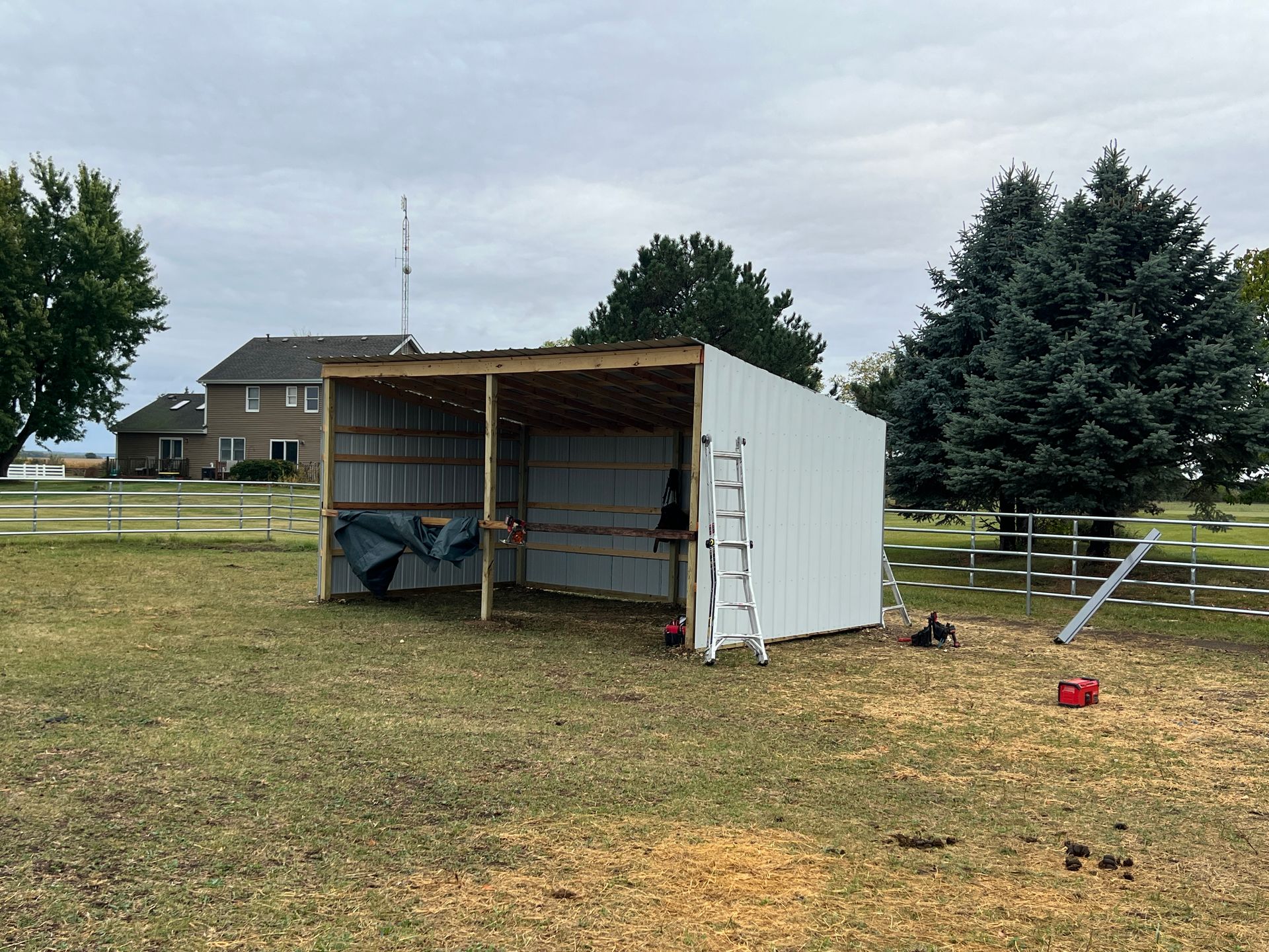 A white, half-finished pole barn structure stands in a grassy field, featuring a ladder and metal siding.