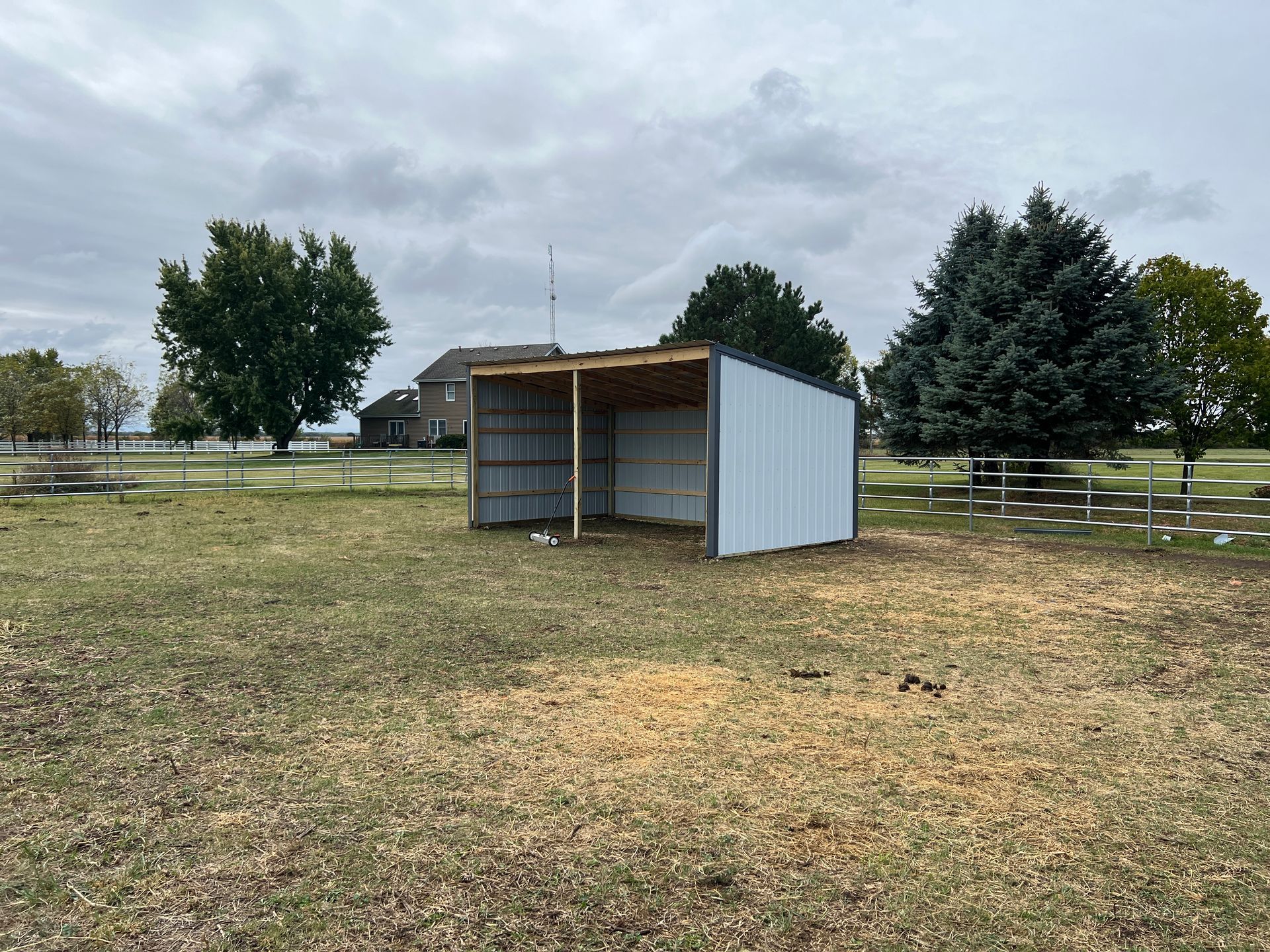 A metal, three-sided run-in shed stands in a grassy, partially shaded pasture on an overcast day.