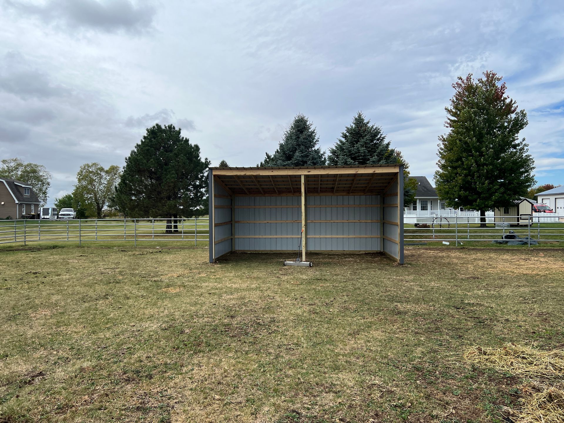 A metal, open-front animal run-in shelter stands in the center of a grassy field under a cloudy sky.
