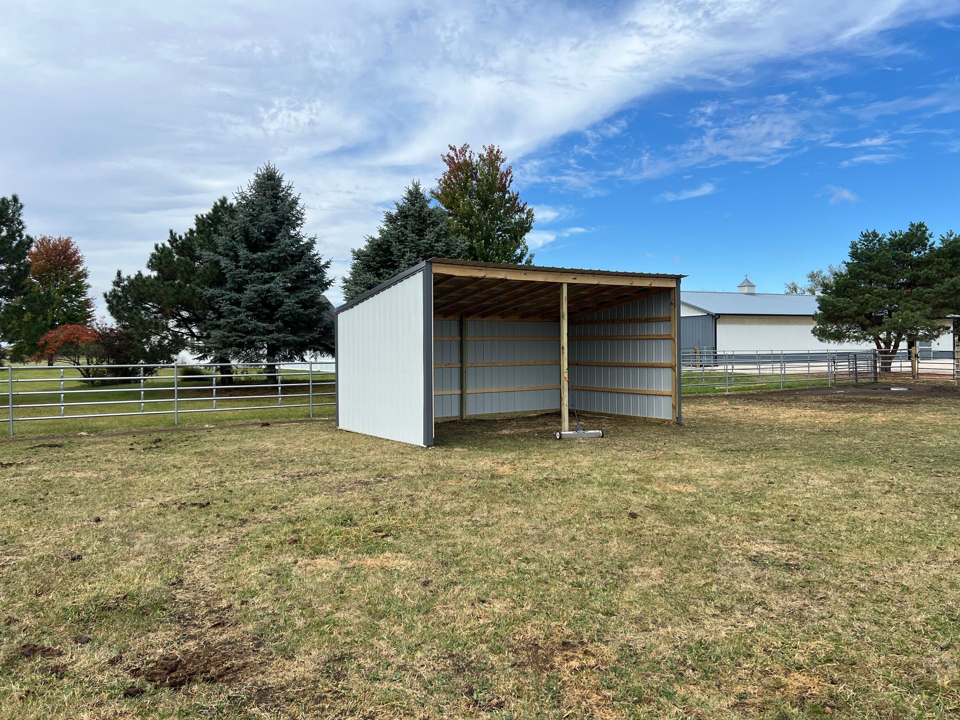 A white, open-front metal shed stands in a grassy field under a partly cloudy sky, with trees in the background.