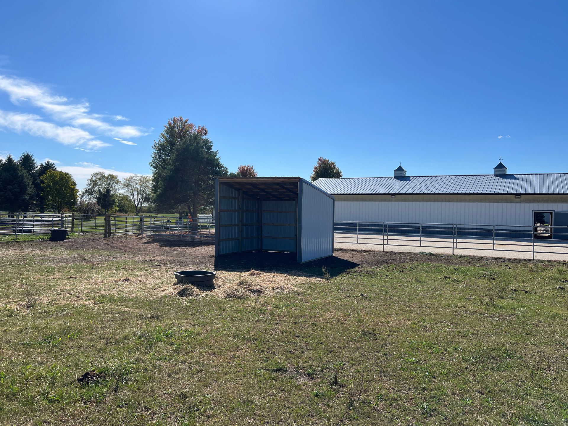 A metal three-sided animal shelter stands in a grass field next to a large white building under a clear blue sky.