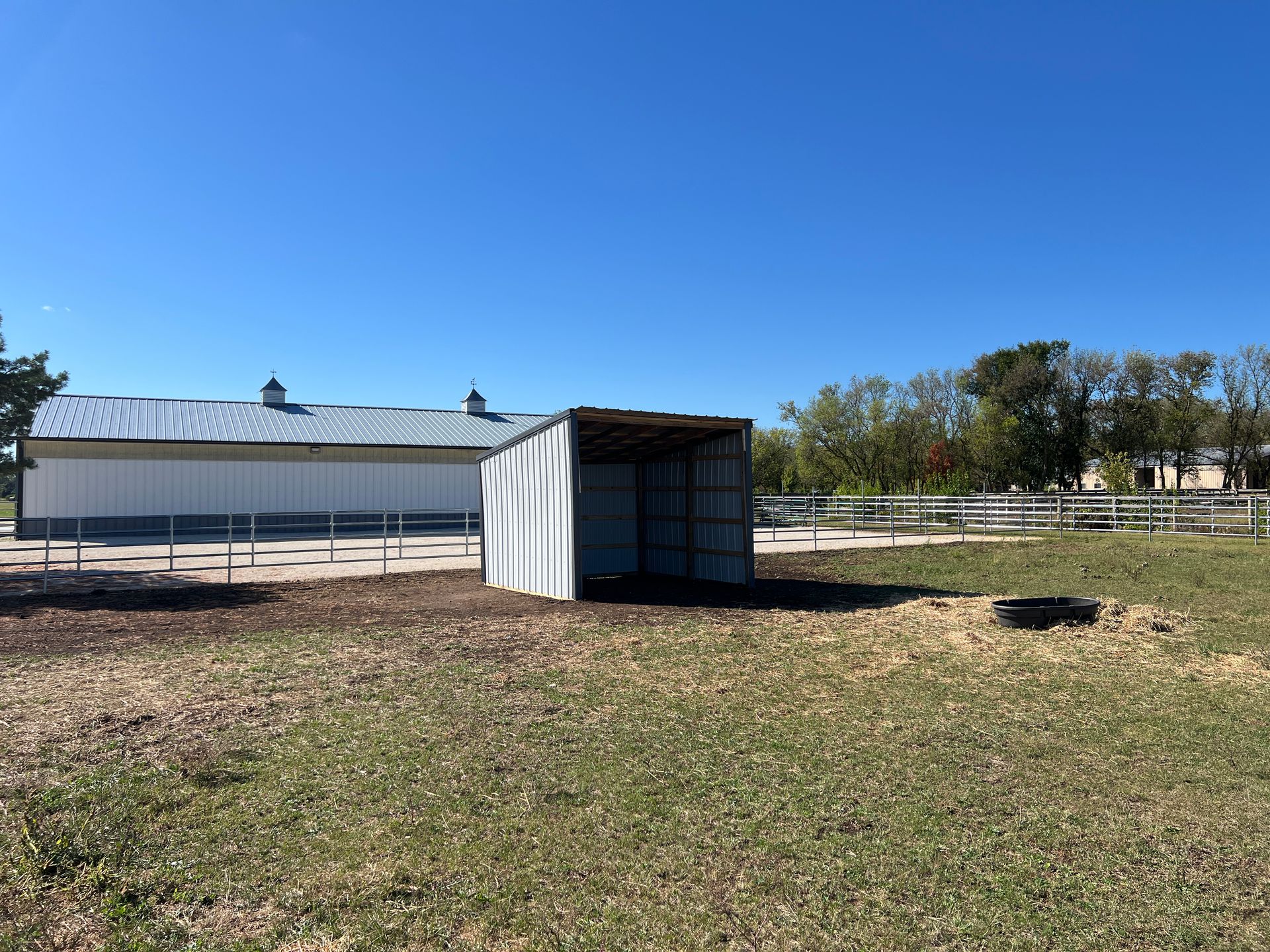 A white shed sits in a grassy field in front of a long, white building under a clear blue sky.