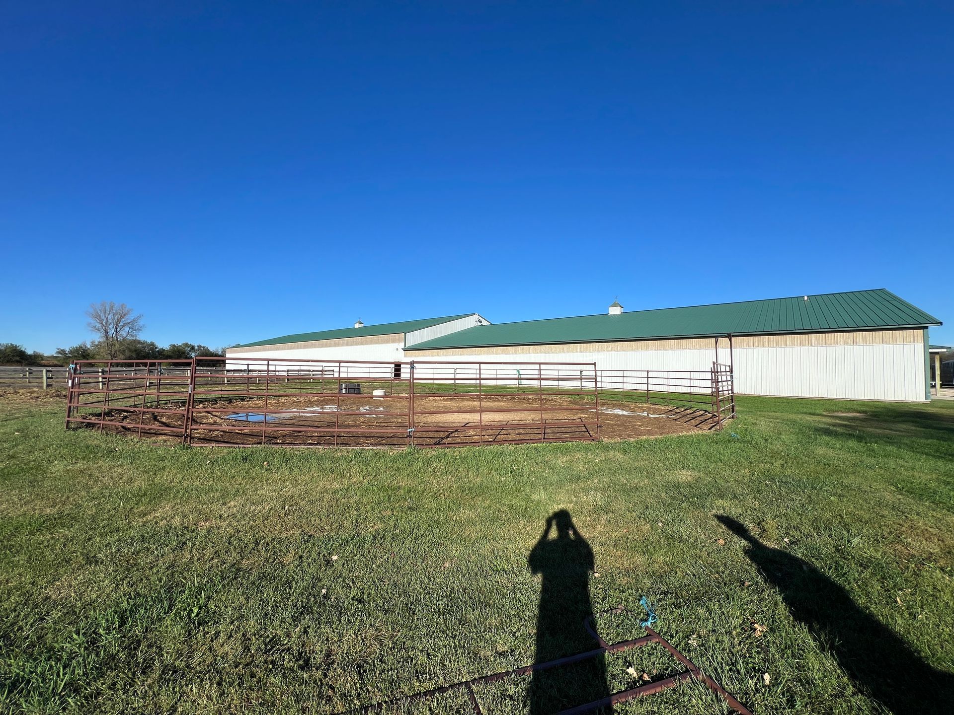 A circular metal round pen sits in a grassy field in front of a long white agricultural building with a green roof.