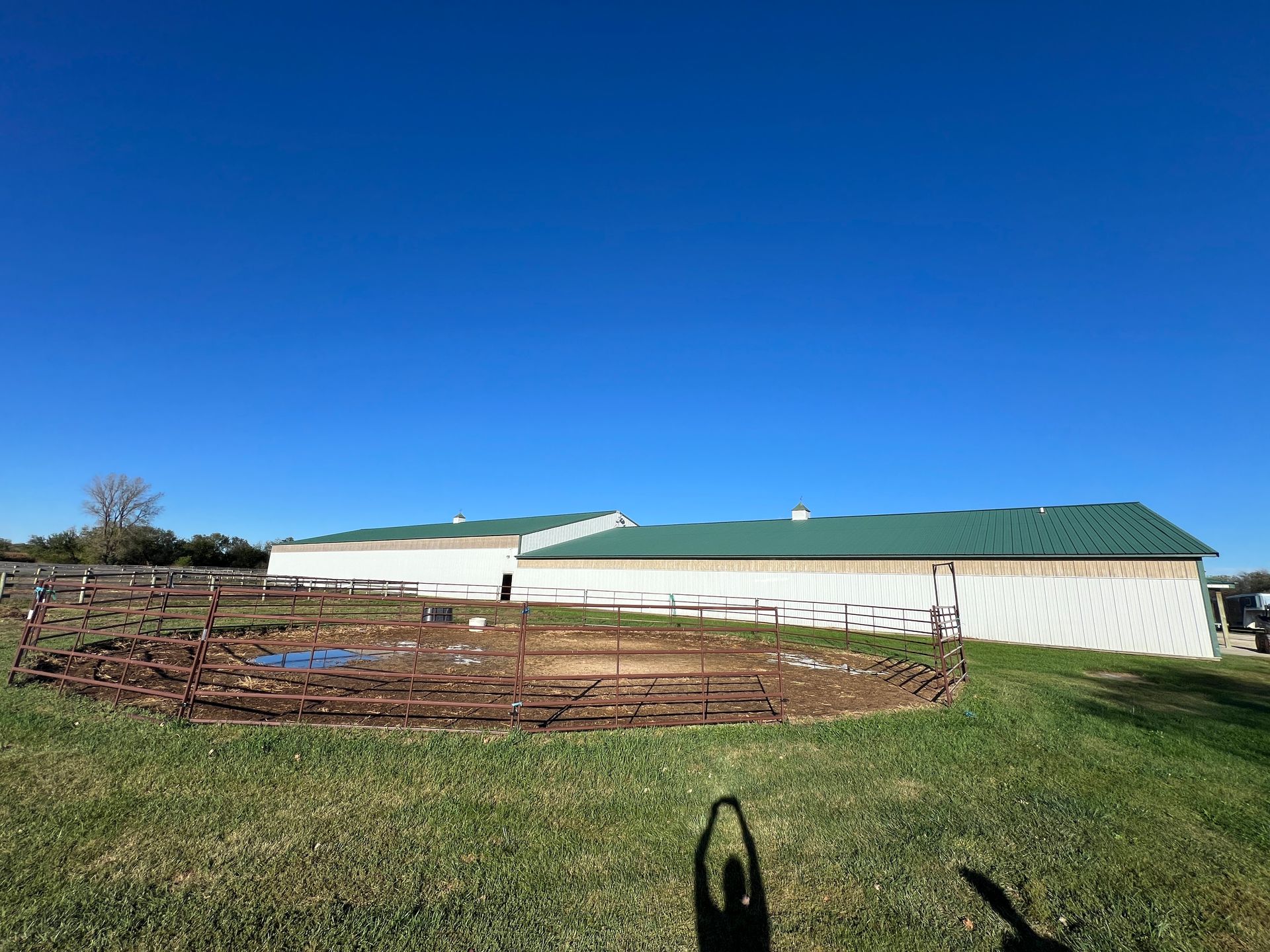 A round metal pen sits in a grassy field in front of a long, white agricultural building with a green metal roof.