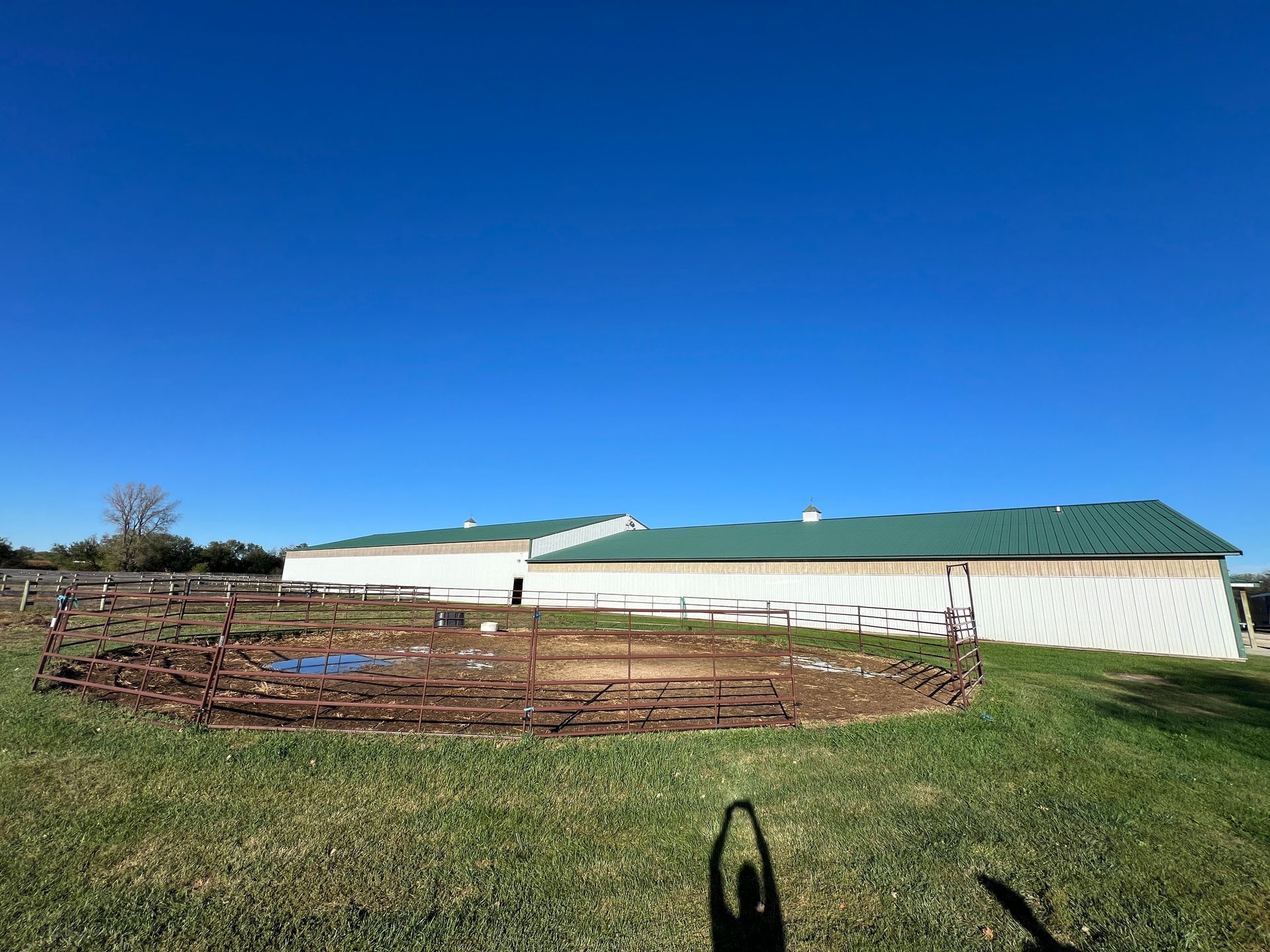 A round metal corral sits in a grassy field in front of a long, white agricultural building with a green roof.