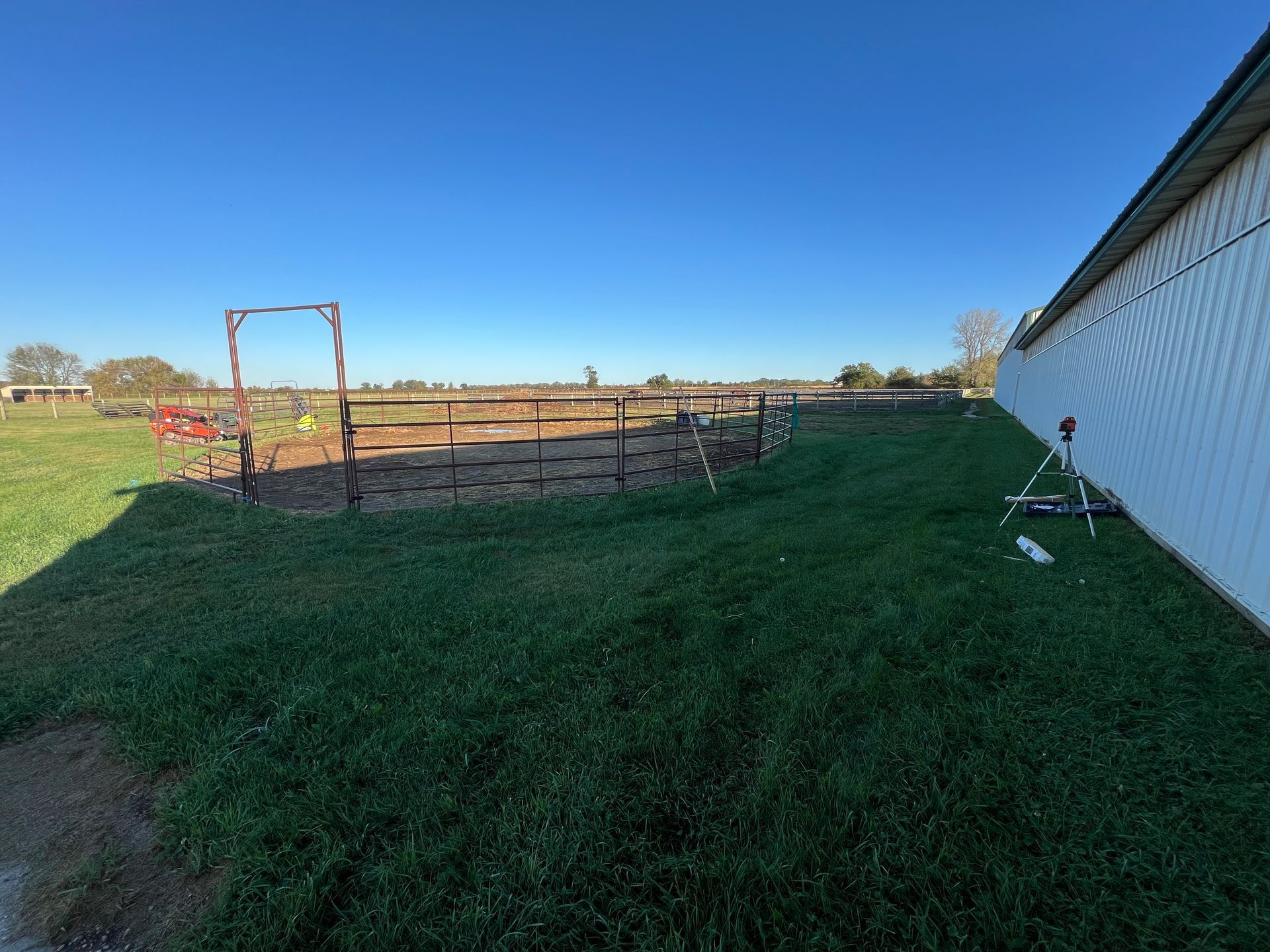 A rural outdoor area with a circular horse training pen, a white building on the right, and a clear blue sky.