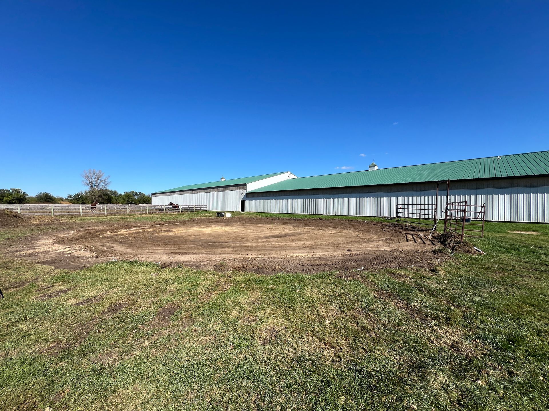 A dirt circle, possibly a round pen, sits in a grassy field in front of a long metal barn under a clear blue sky.