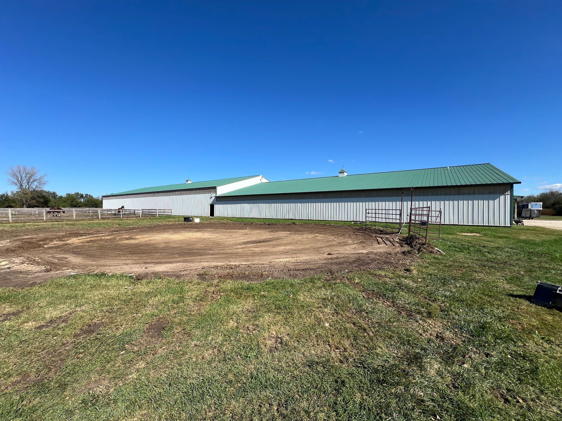 A dirt riding ring in front of a long metal barn with a green roof under a clear blue sky.