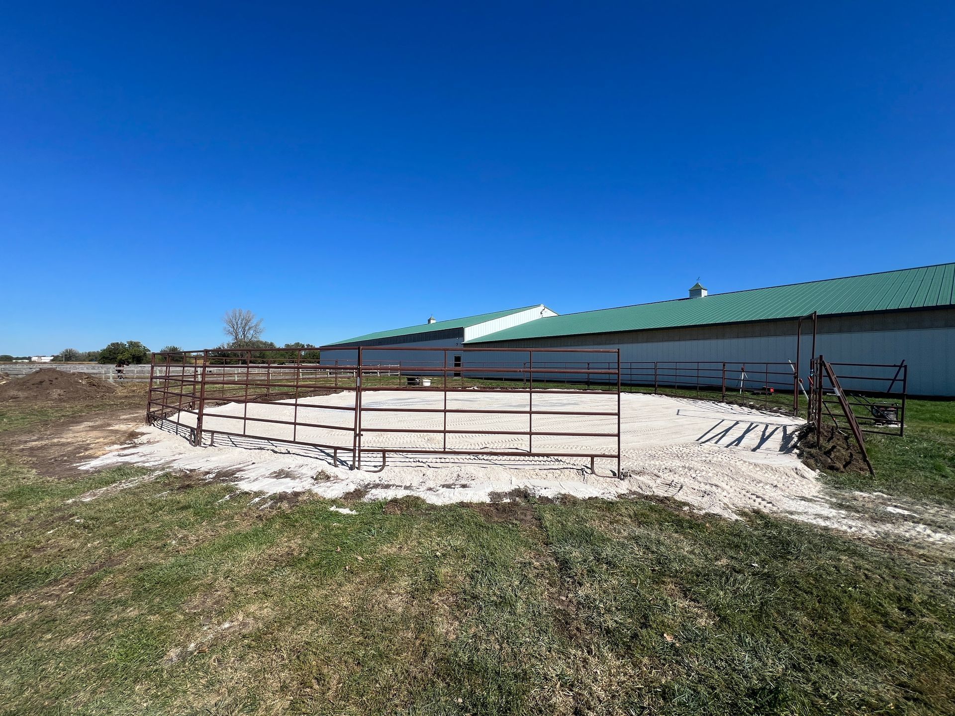 A round training pen with metal railings sits on a bed of white sand in a grassy field next to a large farm building.