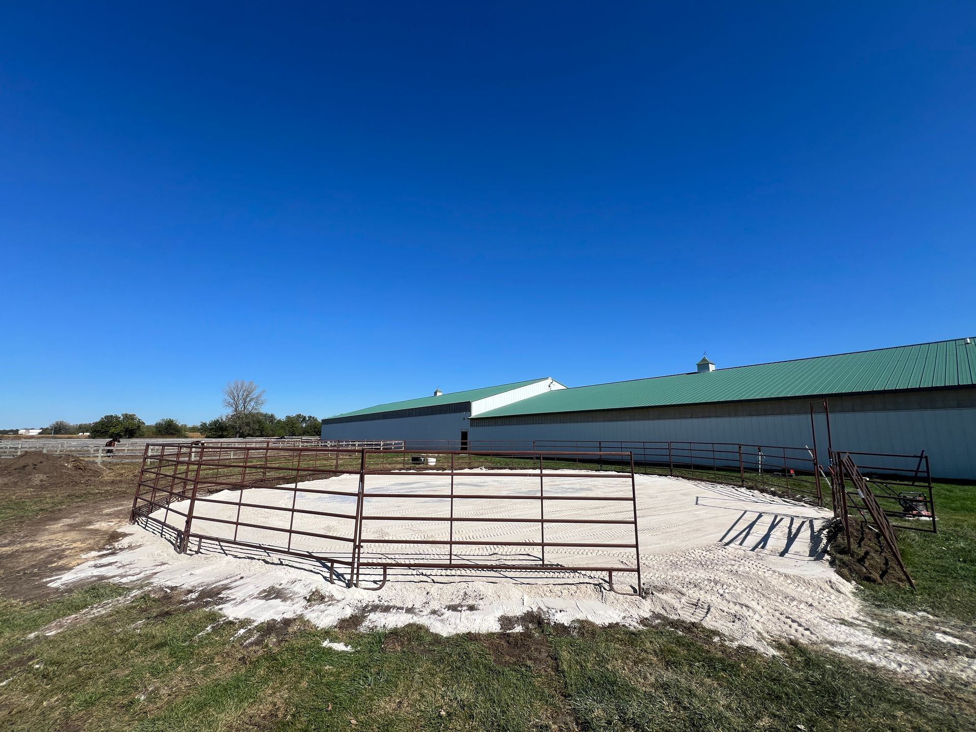 A circular metal pen with a sandy base sits in an open field next to a long, low white building with a green roof.