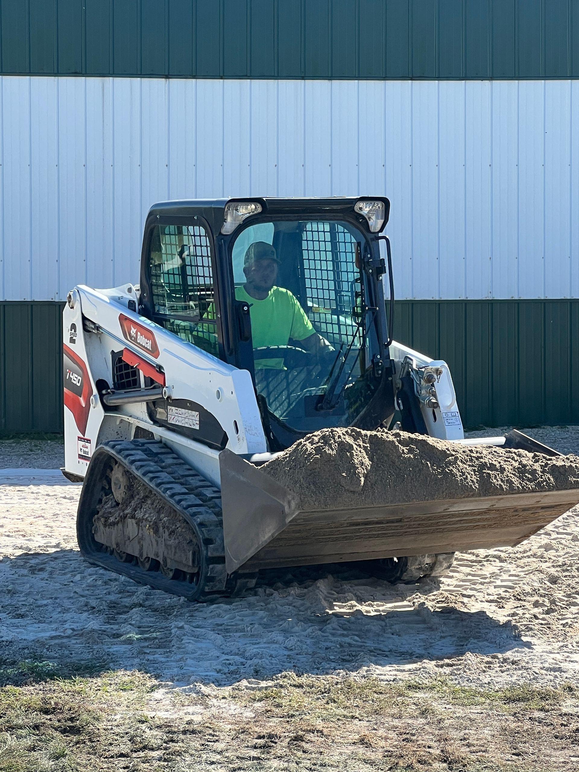 A person wearing a high-visibility green shirt operates a white Bobcat compact track loader carrying a bucket of dirt.