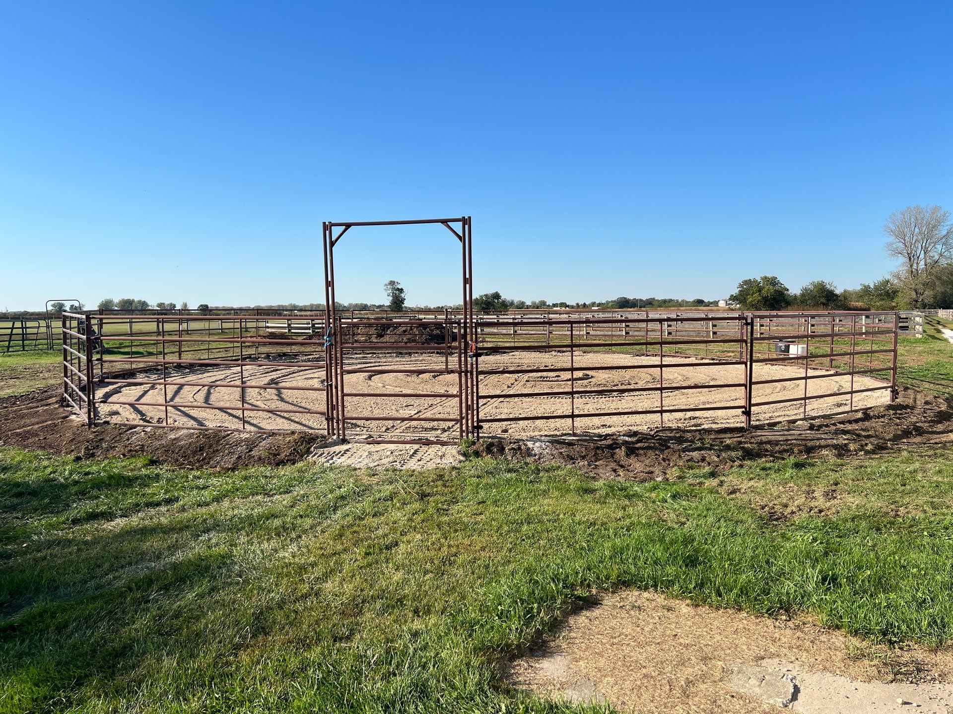 A circular horse training pen with metal fencing and a gated entrance, set in a grassy outdoor field under a clear sky.