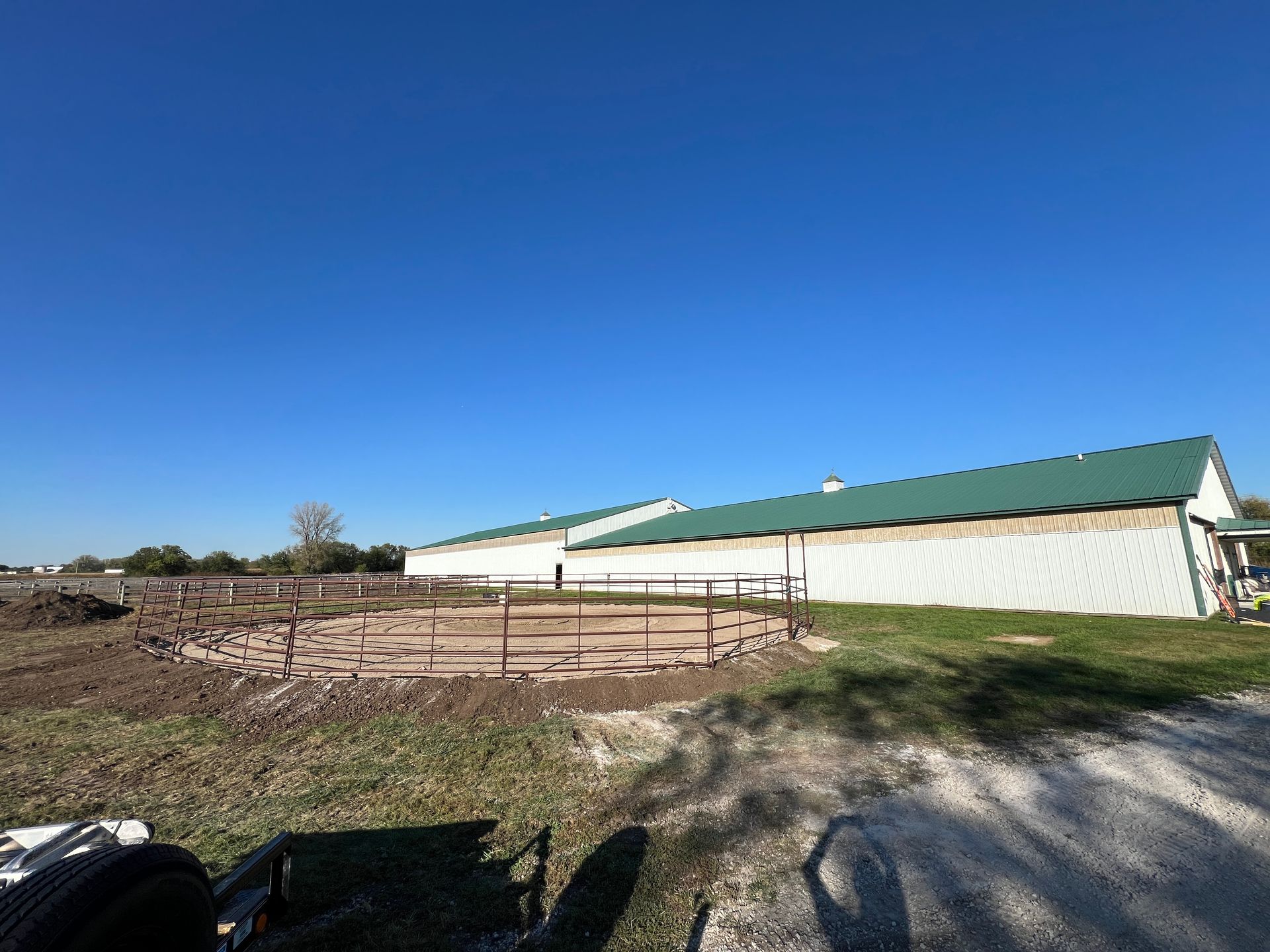 A round metal horse pen sits in a dirt field next to a long agricultural building with a green roof under a blue sky.