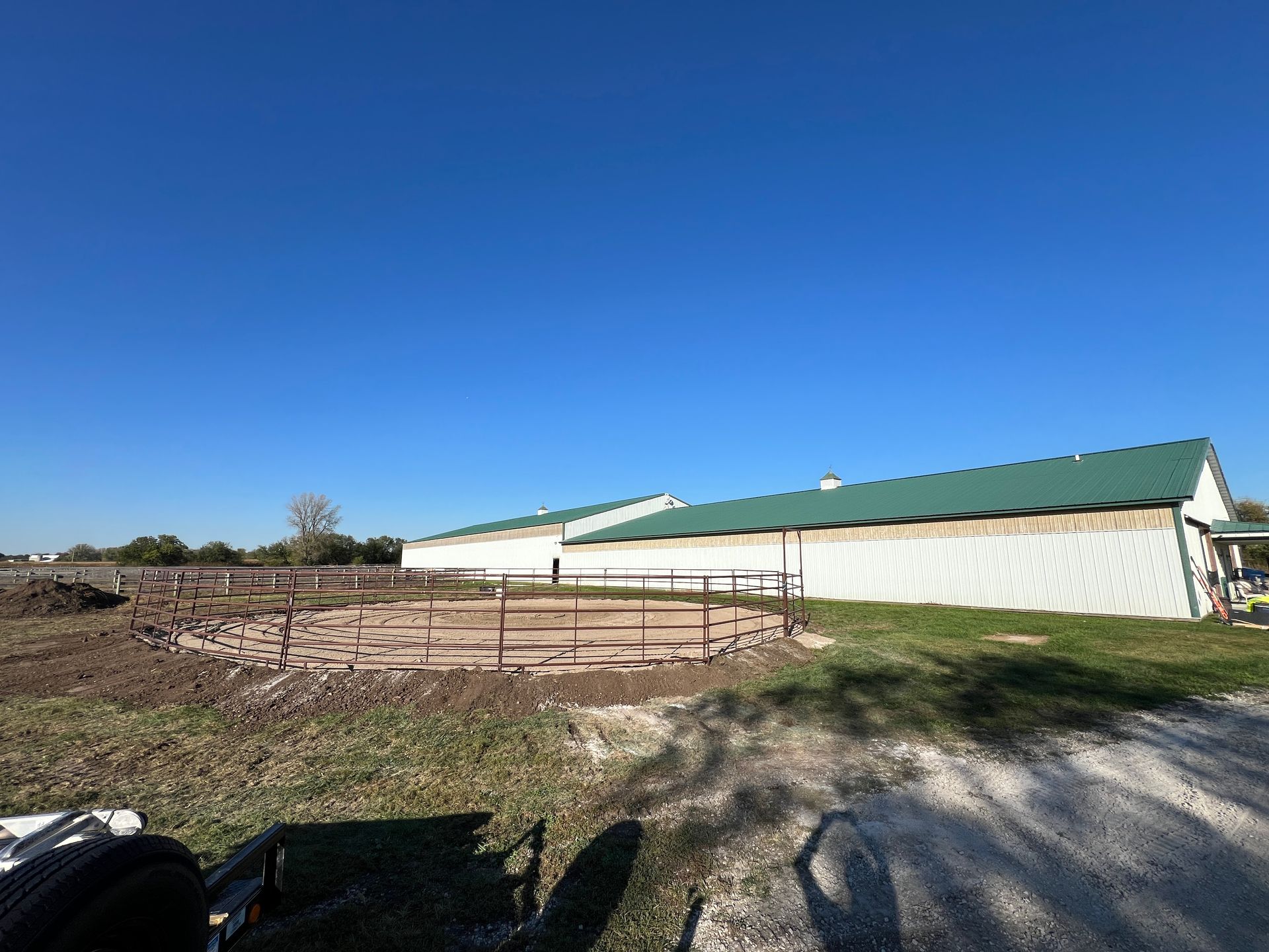 A circular metal pen sits in a dirt field in front of a long, white agricultural building with a green roof.