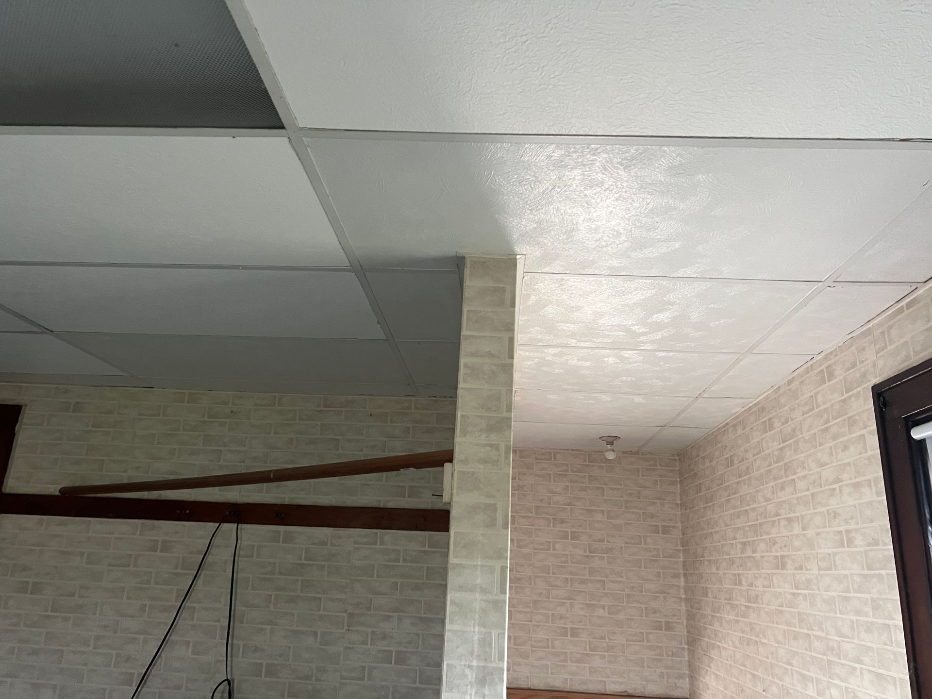 A partially finished room with a drop ceiling, beige brick-patterned wallpaper, and an exposed wooden ceiling joist.