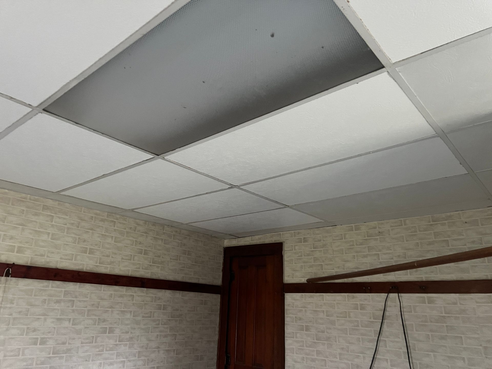 A drop ceiling with a recessed rectangular light fixture, beige wallpaper, and horizontal wooden trim in a room.