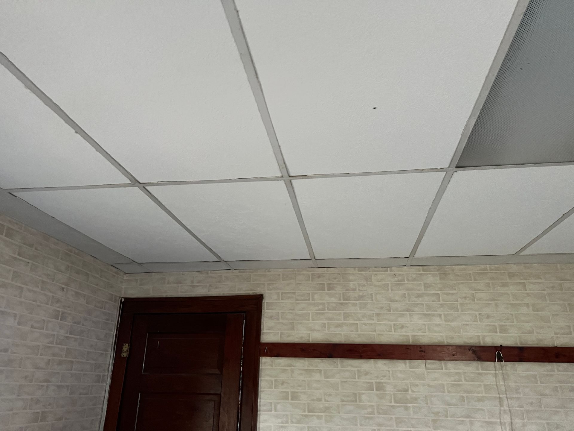 White drop ceiling panels installed above a room with patterned wallpaper and a dark wooden door frame.