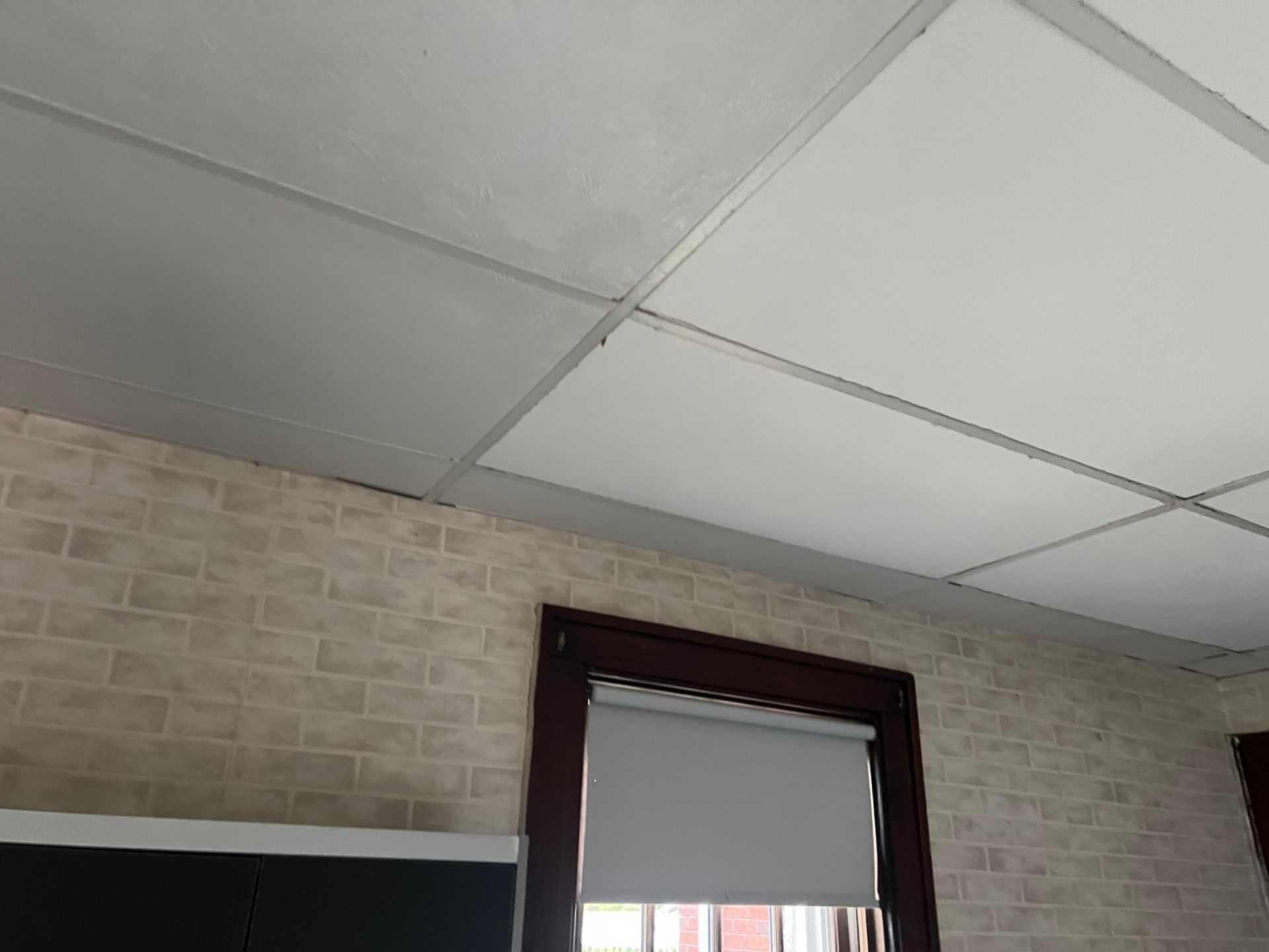 A low-angle view of a drop ceiling with white acoustic tiles, positioned above walls covered in light brick wallpaper.