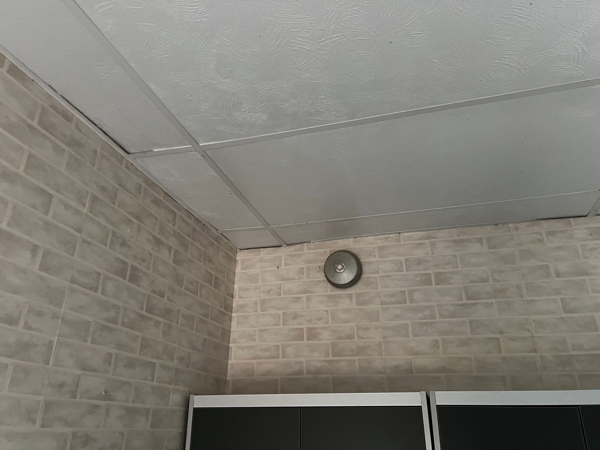 A drop ceiling above a wall covered in light-colored faux brick wallpaper, with a small circular vent mounted on the wall.