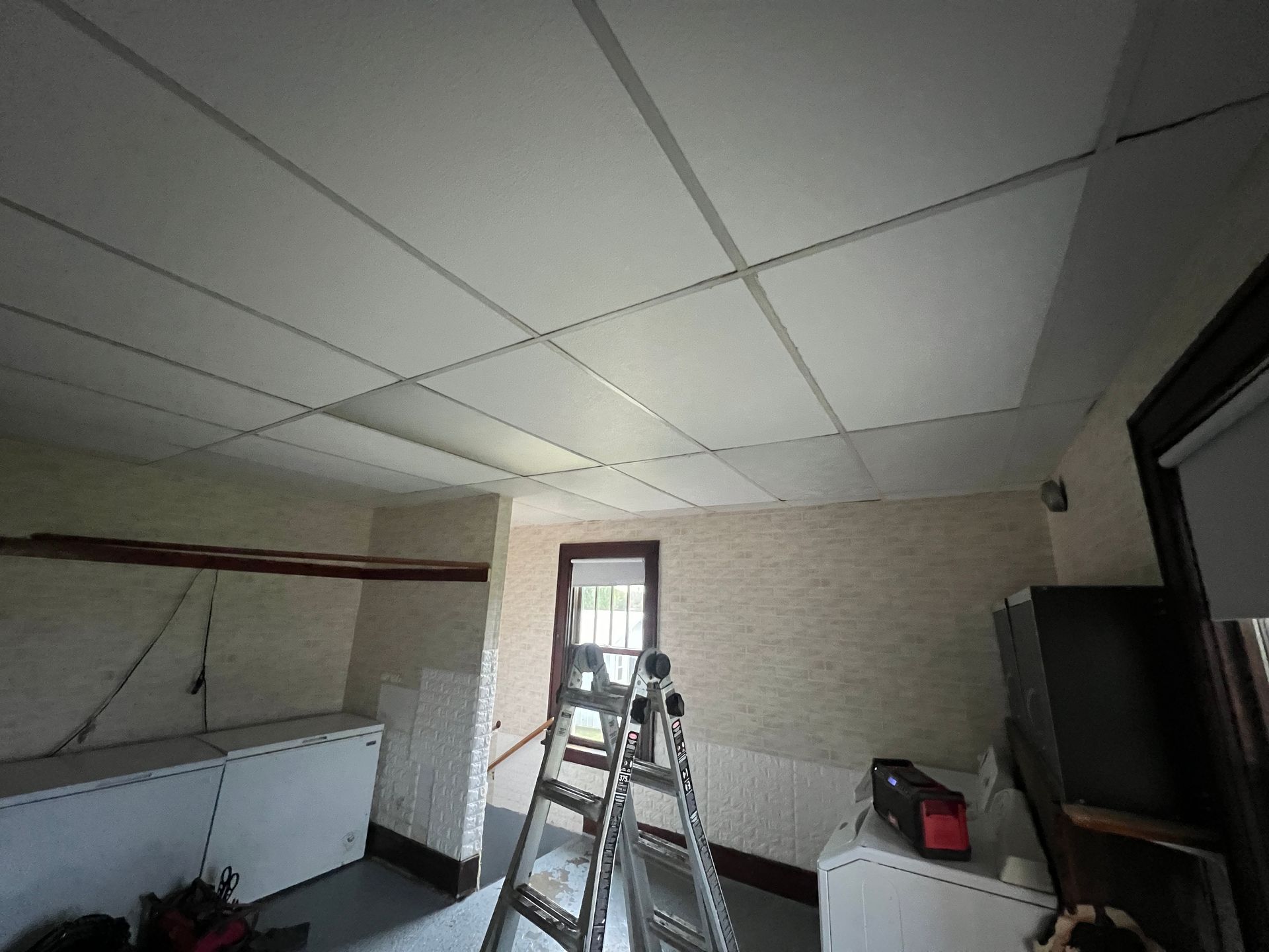 A room with a white drop ceiling, cream walls, a silver stepladder in the center, a chest freezer, and laundry equipment.