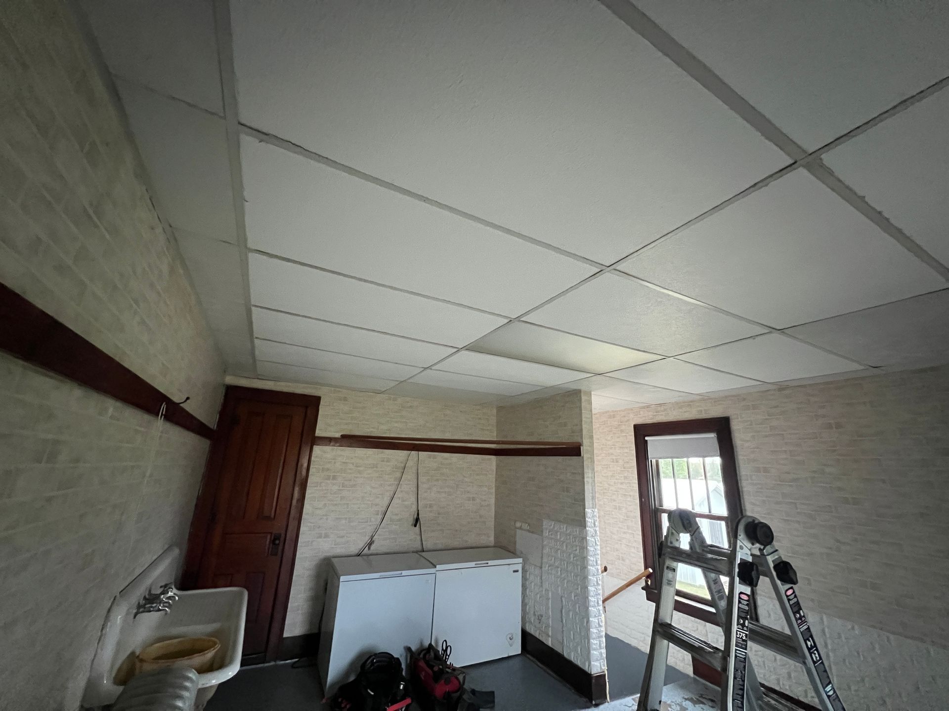 A laundry room with a drop ceiling, white appliances, a sink, and a step ladder, under renovation.