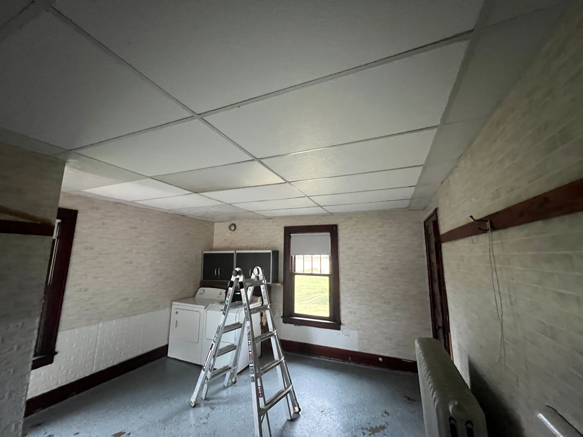 A room undergoing renovation with a stepladder, a washing machine, white brick walls, and a drop ceiling.