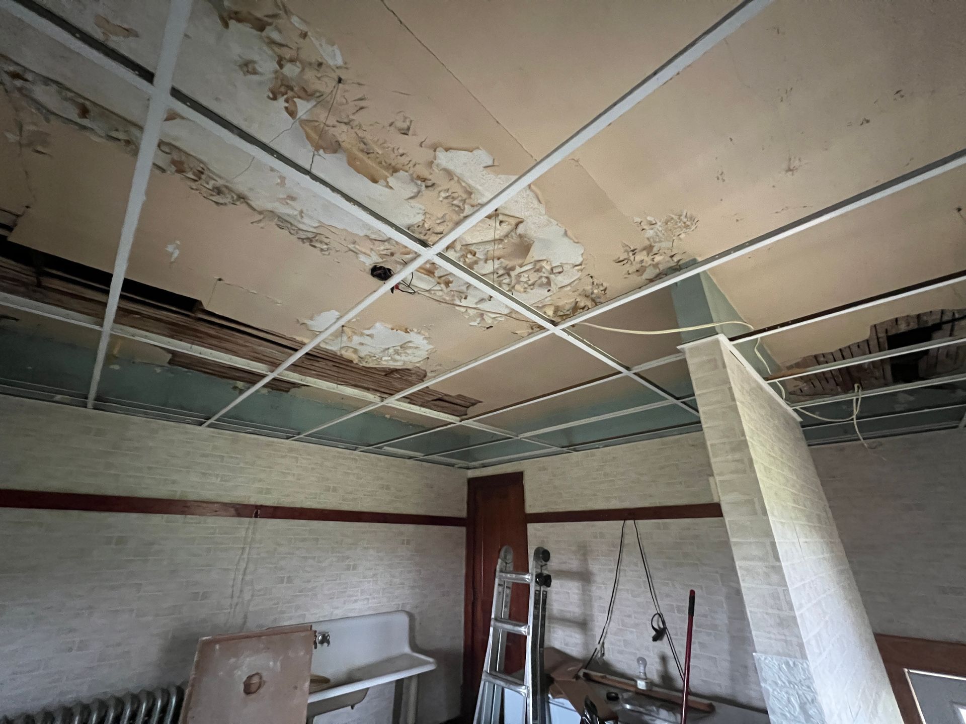 A room under renovation with a damaged ceiling, exposed metal grid, a ladder, and a wall-mounted sink.