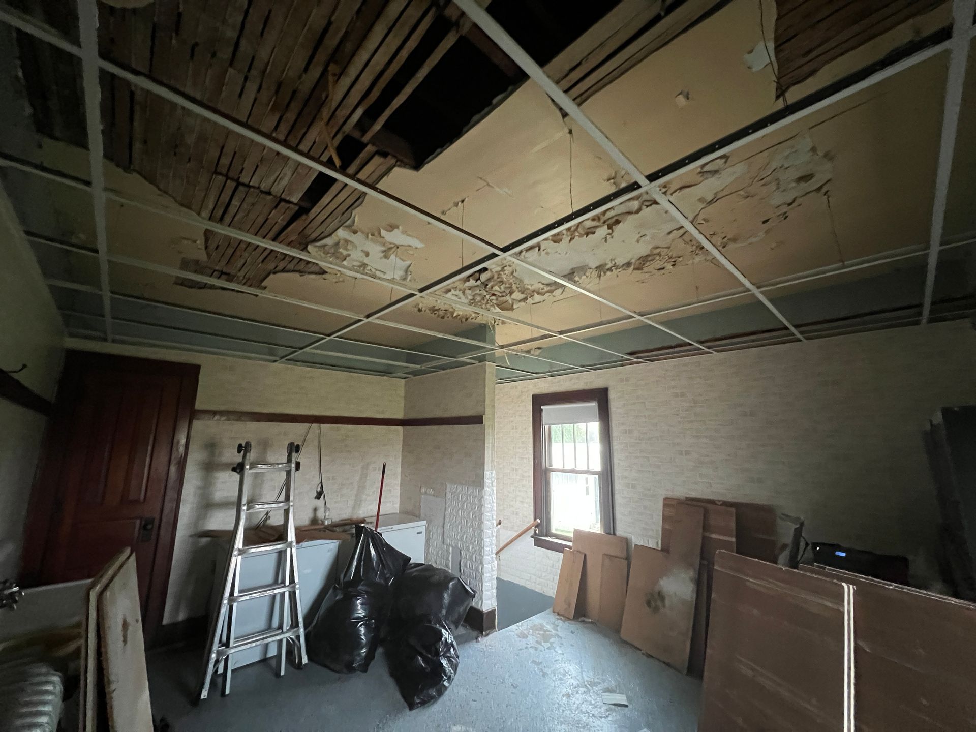 A room undergoing renovation with exposed lath ceilings, a metal grid frame, a metal ladder, and debris in black bags.