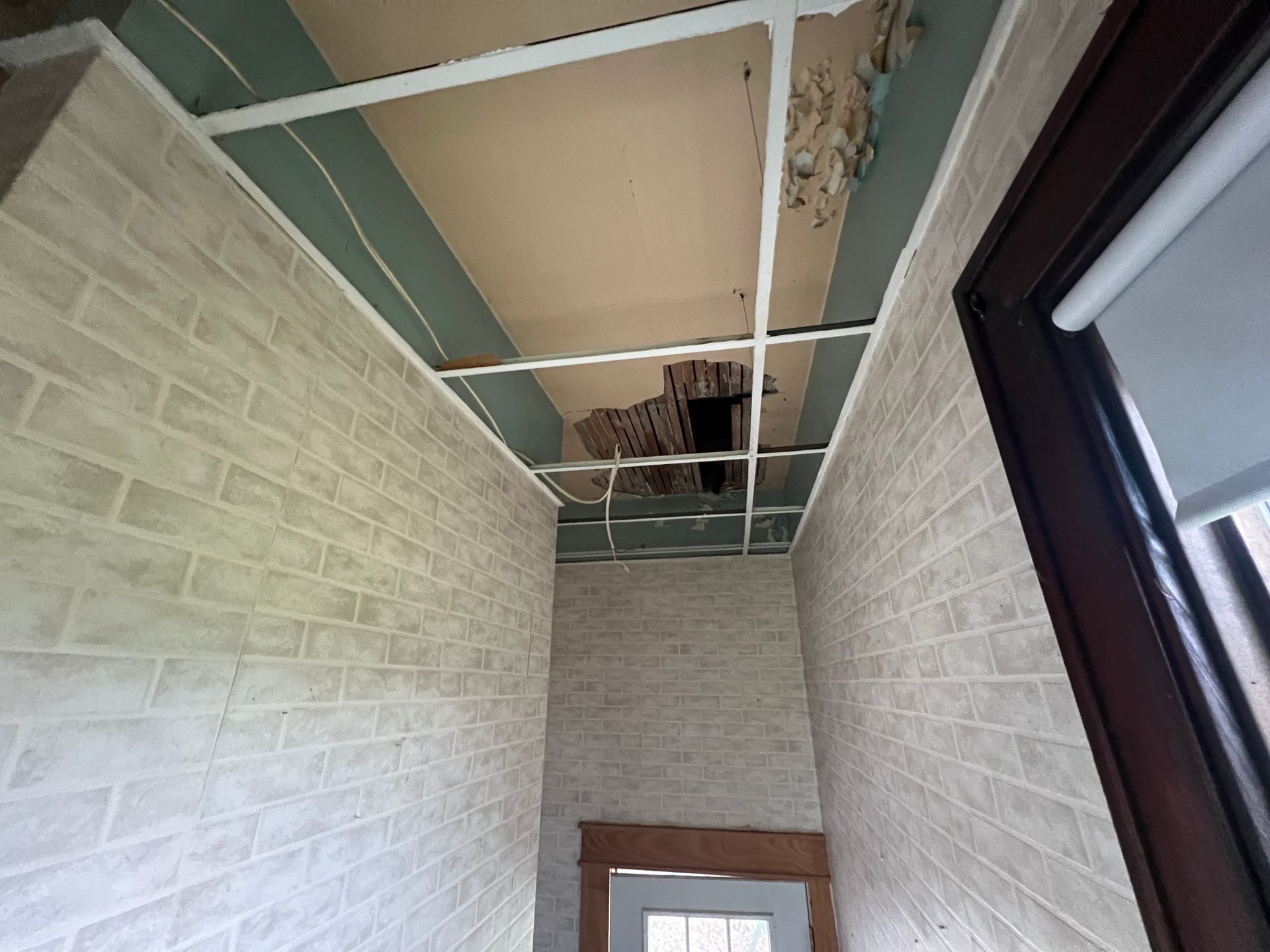An interior hallway with brick-patterned wallpaper, a drop ceiling grid, and a hole exposing the sub-ceiling structure.