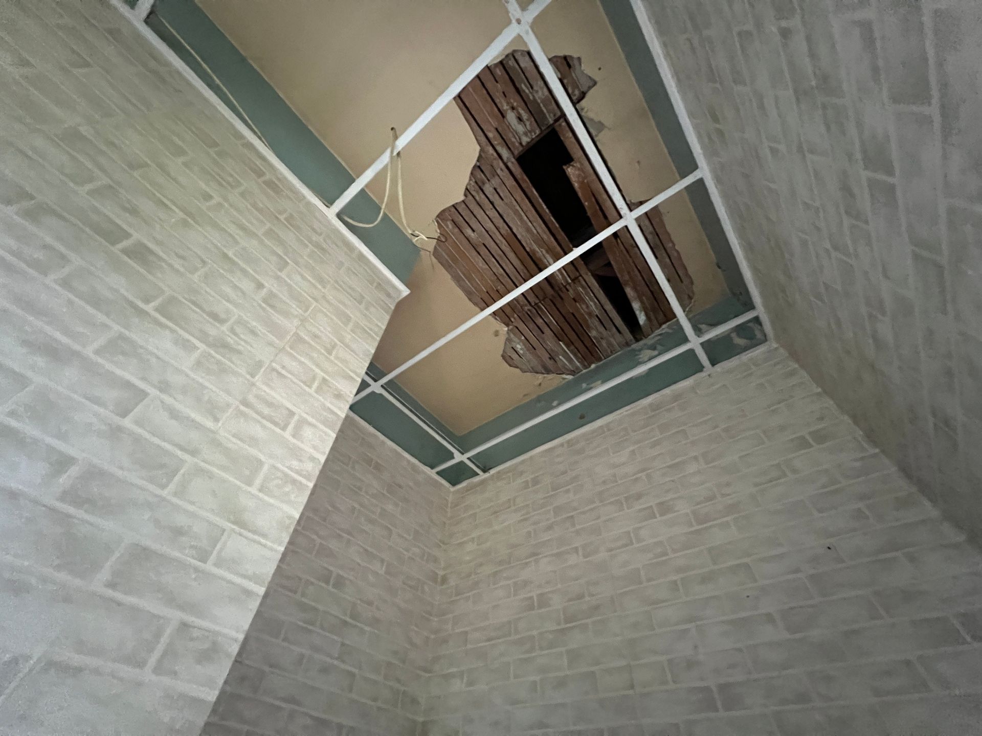 A damaged ceiling with a large hole revealing wooden laths, set in a room with light-colored brick-patterned walls.